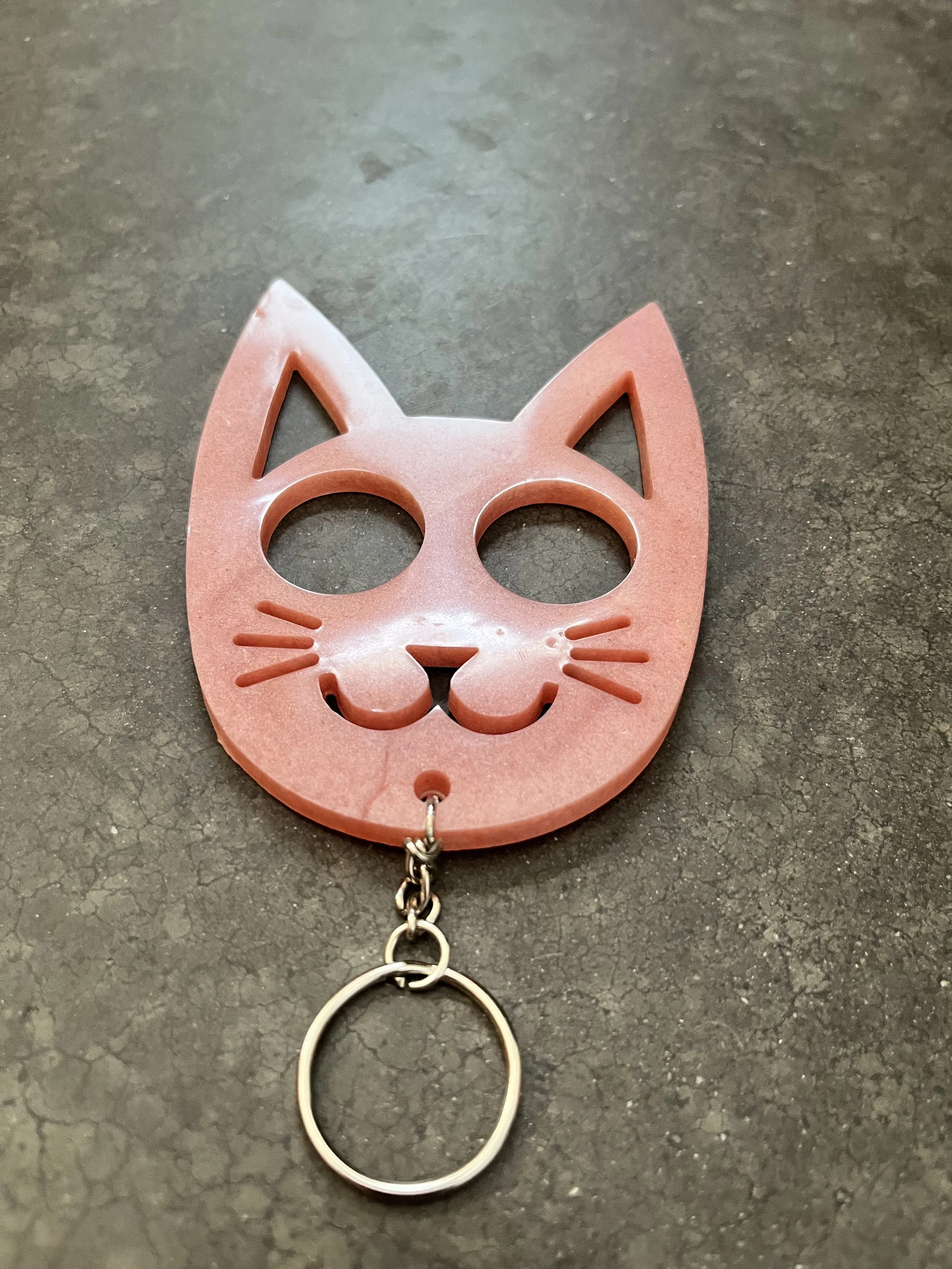 A pink cat-shaped keychain with cutouts for the eyes, nose, mouth, and ears, attached to a metal keyring, on a dark textured surface.