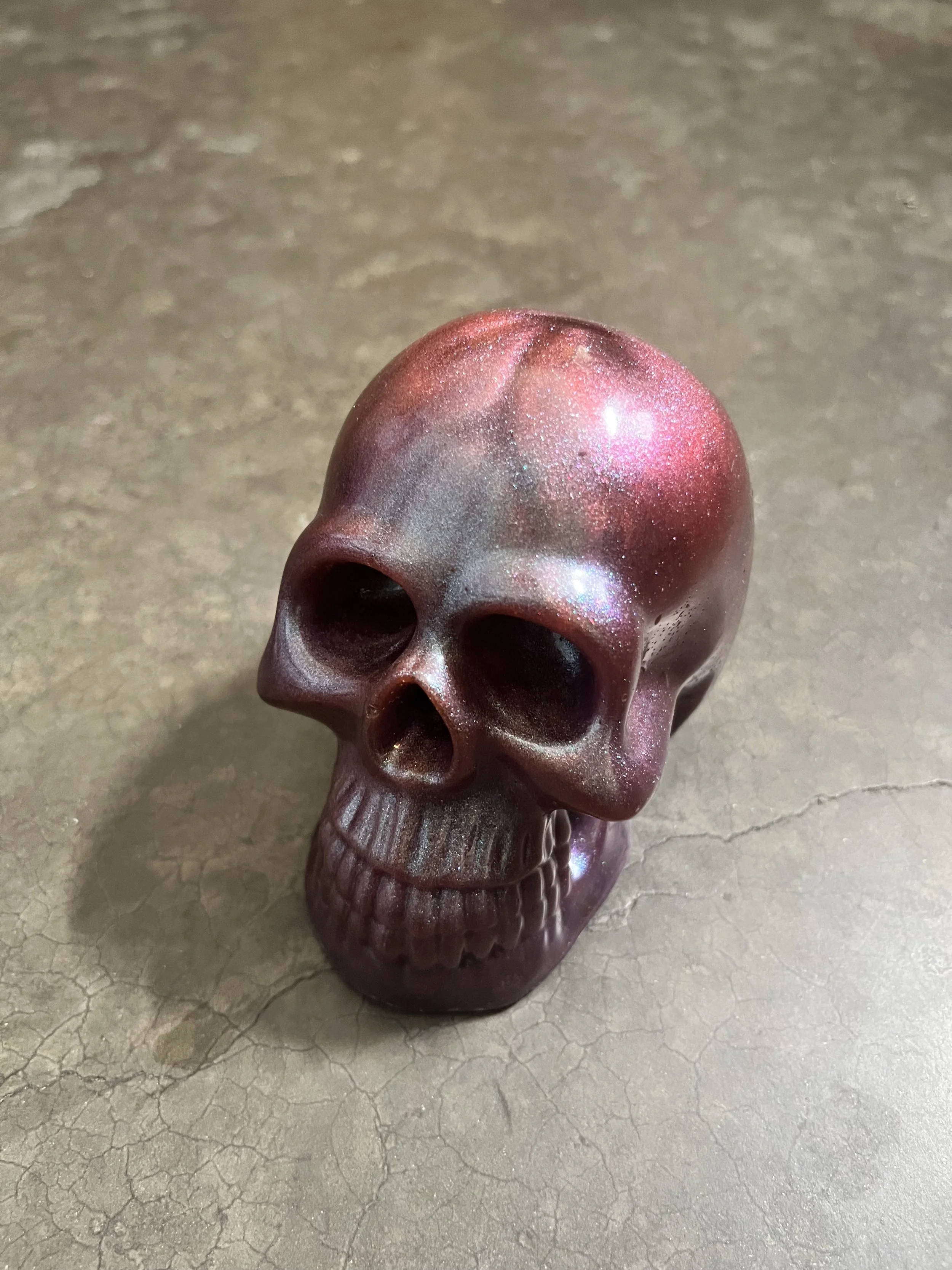 A handmade epoxy resin skull ornament with a shiny, glittery finish, placed on a cracked concrete surface.