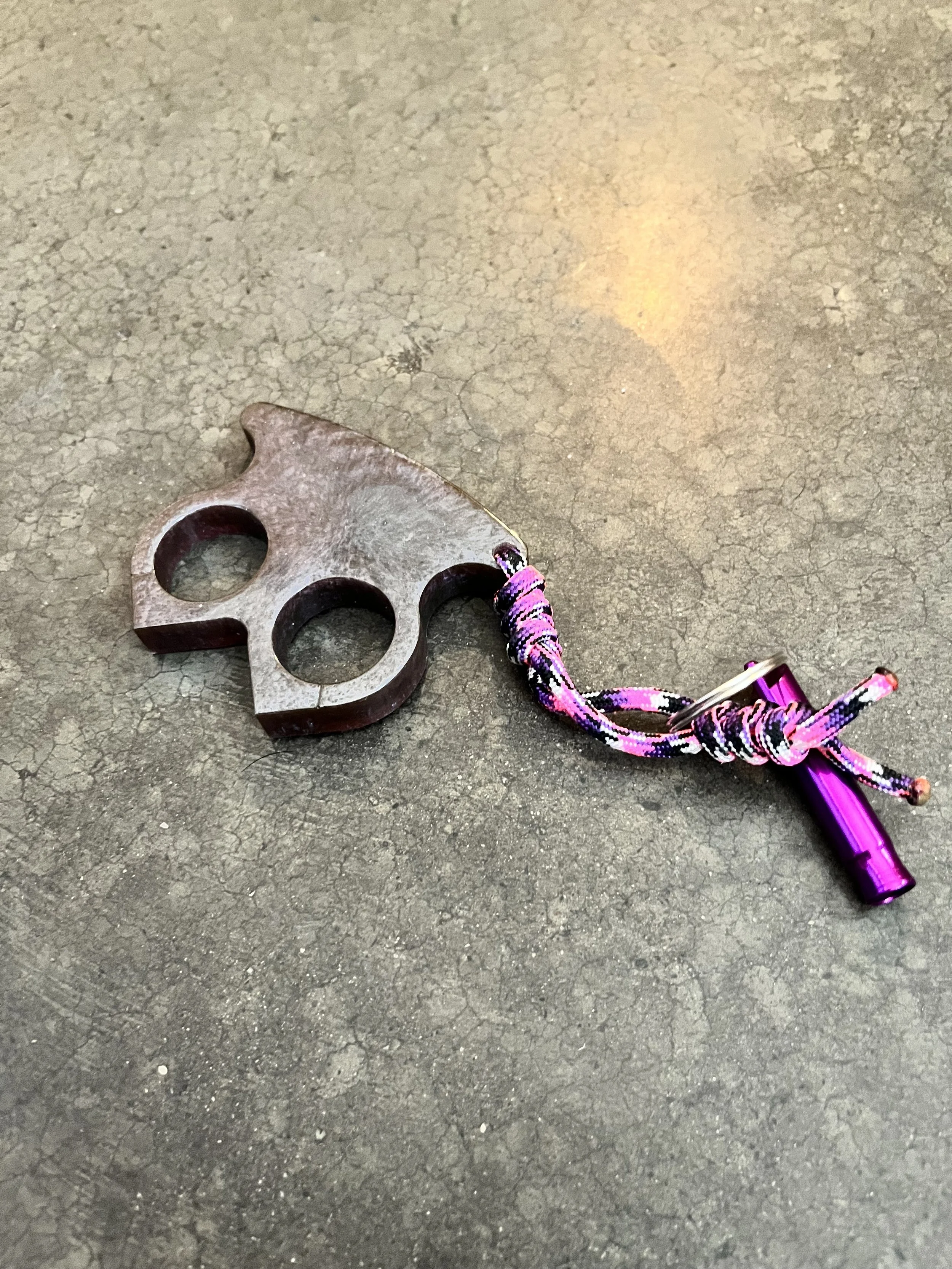 A metal finger graffiti lock and a pink and black braided lanyard with a purple flashlight attached are lying on a rough, cracked asphalt surface.