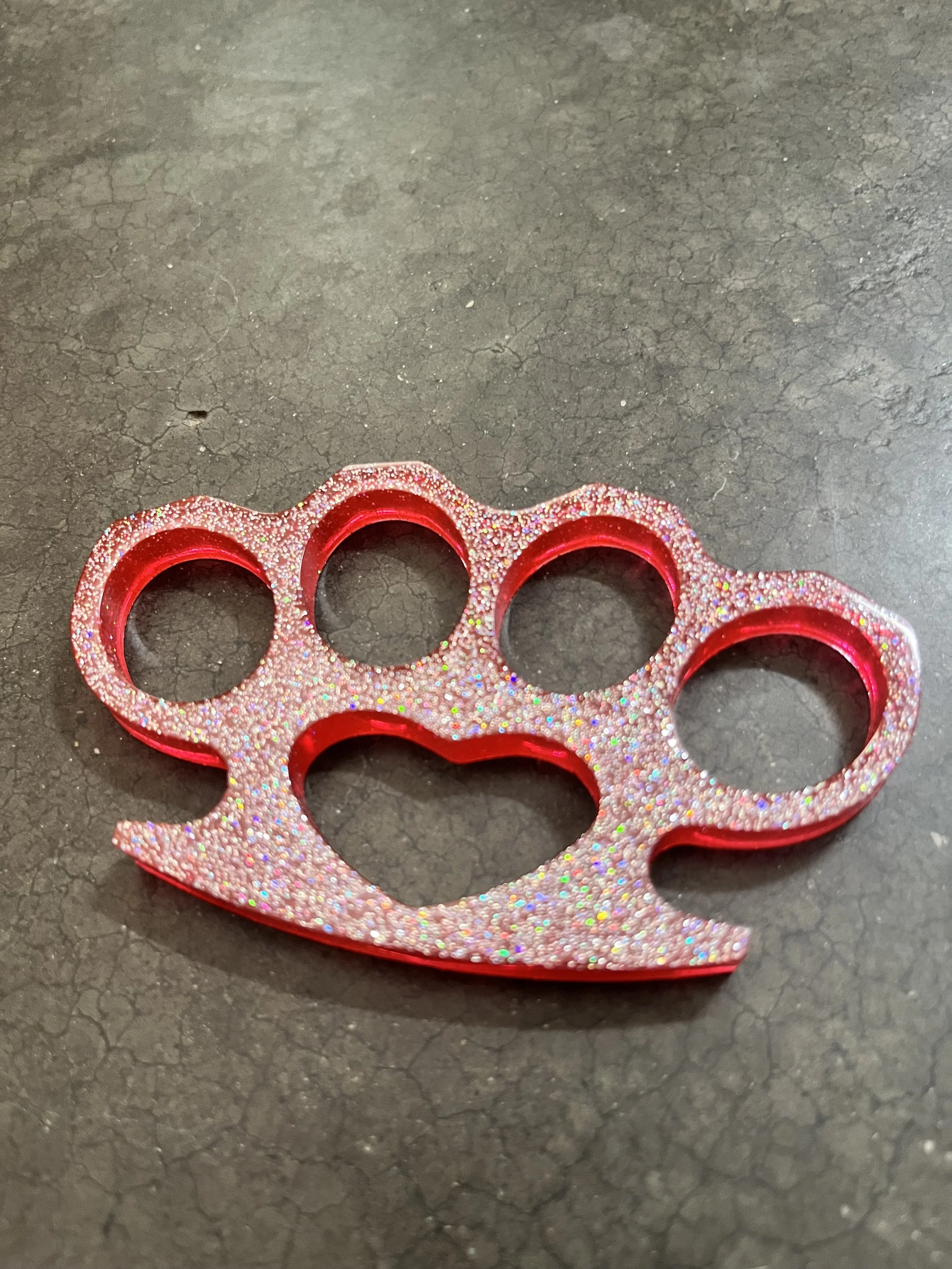 A red and pink glittery brass knuckle keychain with five finger openings on a dark textured surface.
