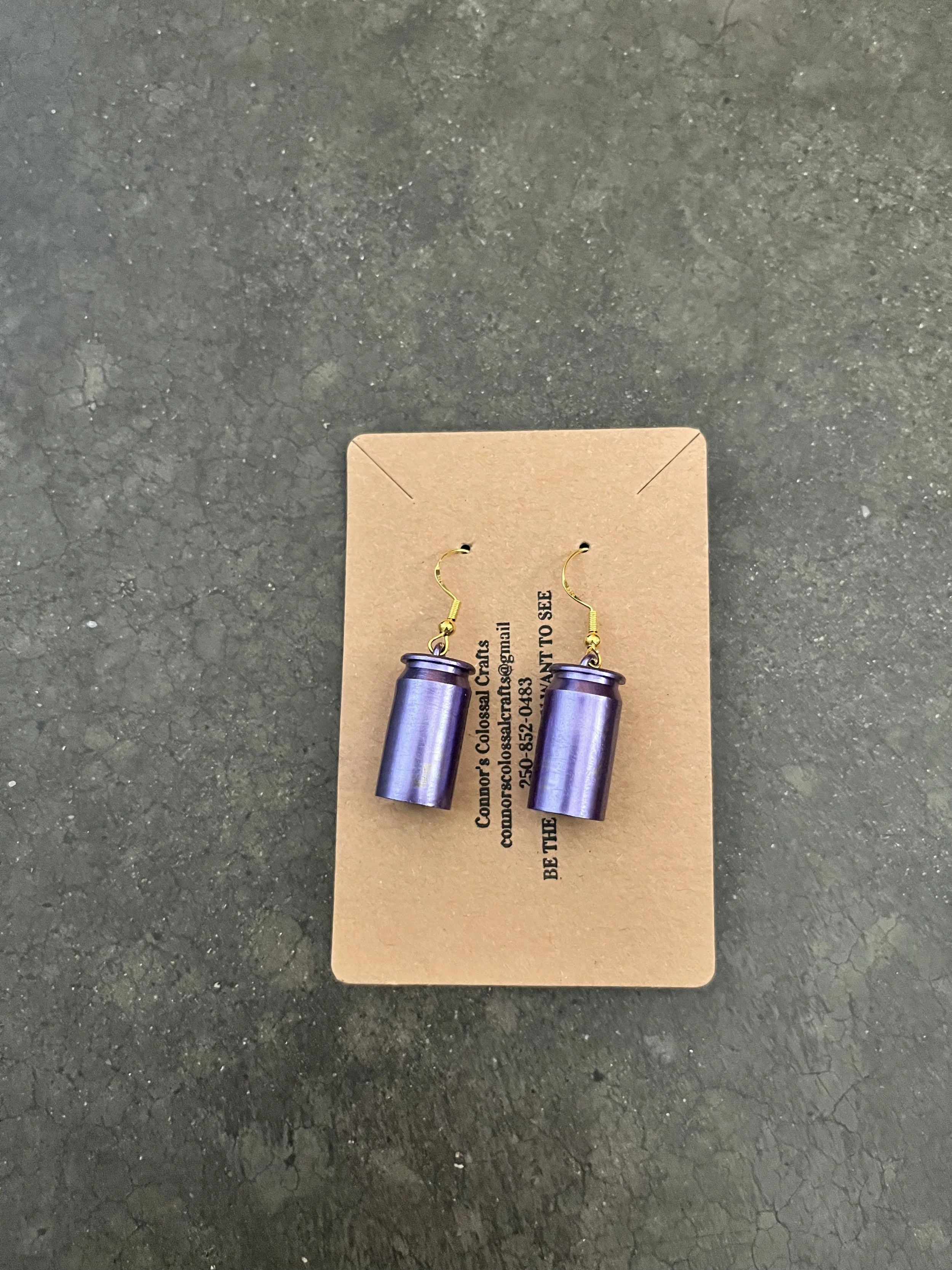 A pair of purple metallic gun casing earrings on a cardboard backing. The backing has text with contact details.