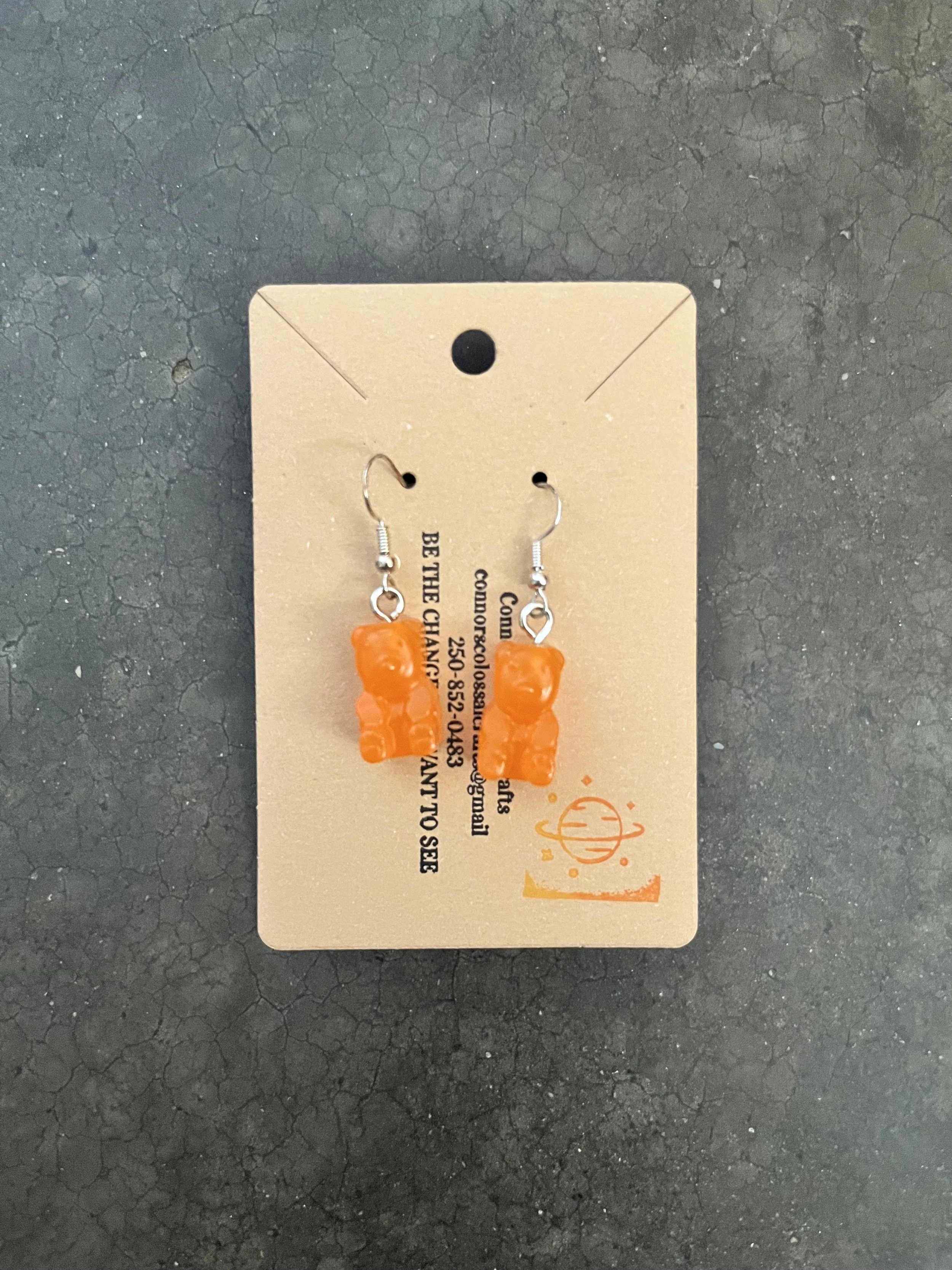 Orange gummy bear earrings on a beige card with black text, placed on a textured black surface.