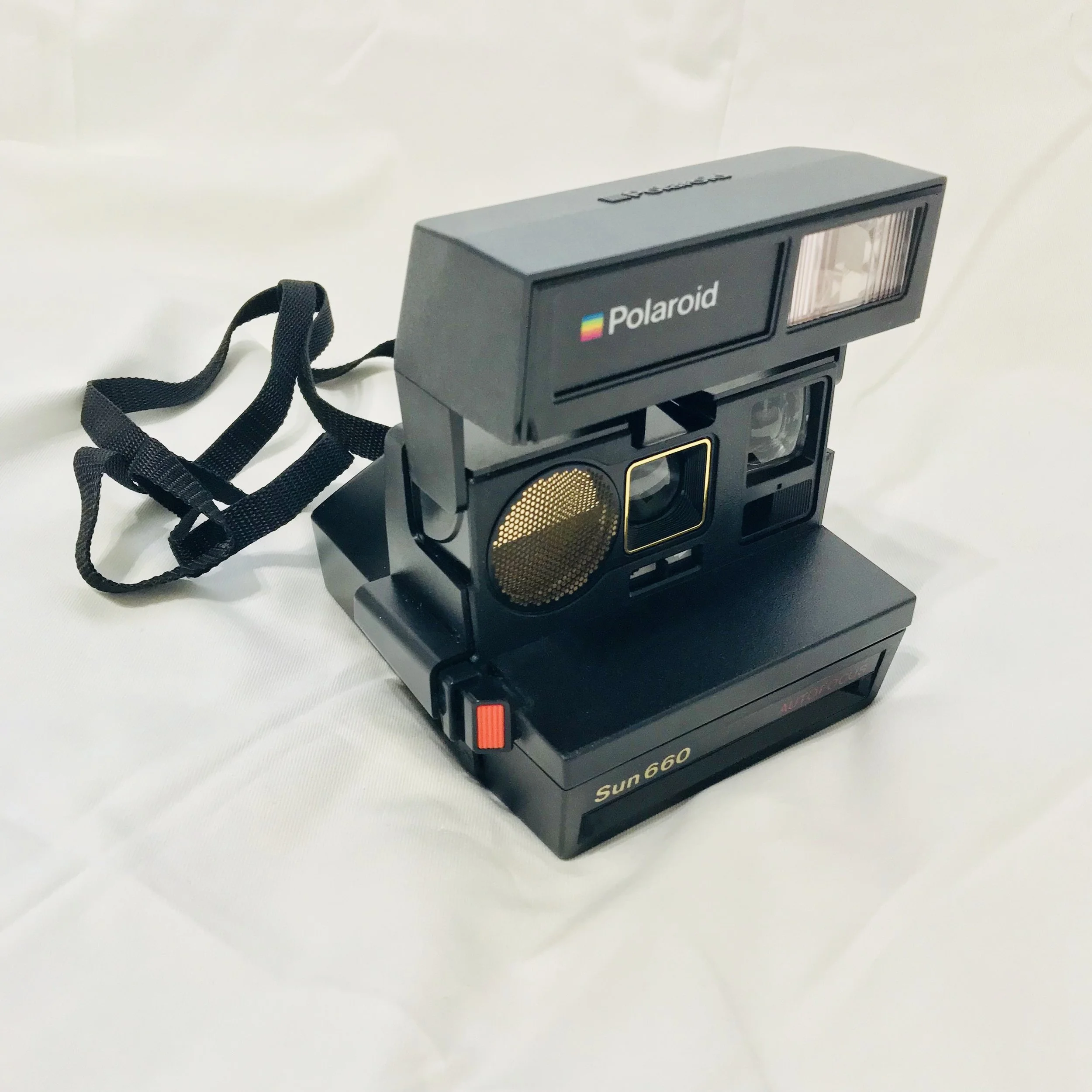 A vintage Polaroid Sun 660 instant film camera with a black body, gold accents, and a strap, placed on a white surface.