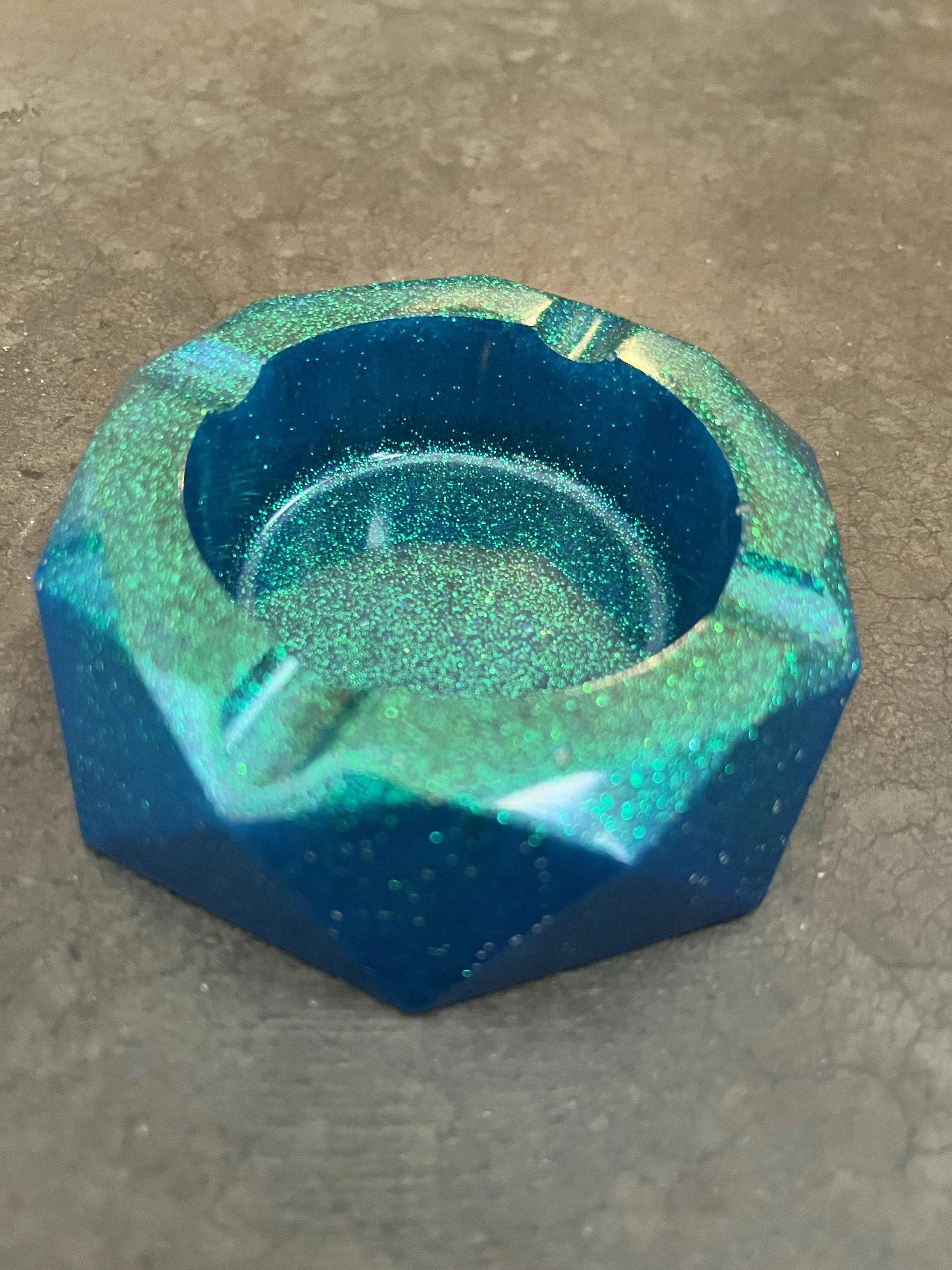 A blue glass hexagonal-shaped ashtray/container with a sparkly, glittery interior placed on a gray surface.