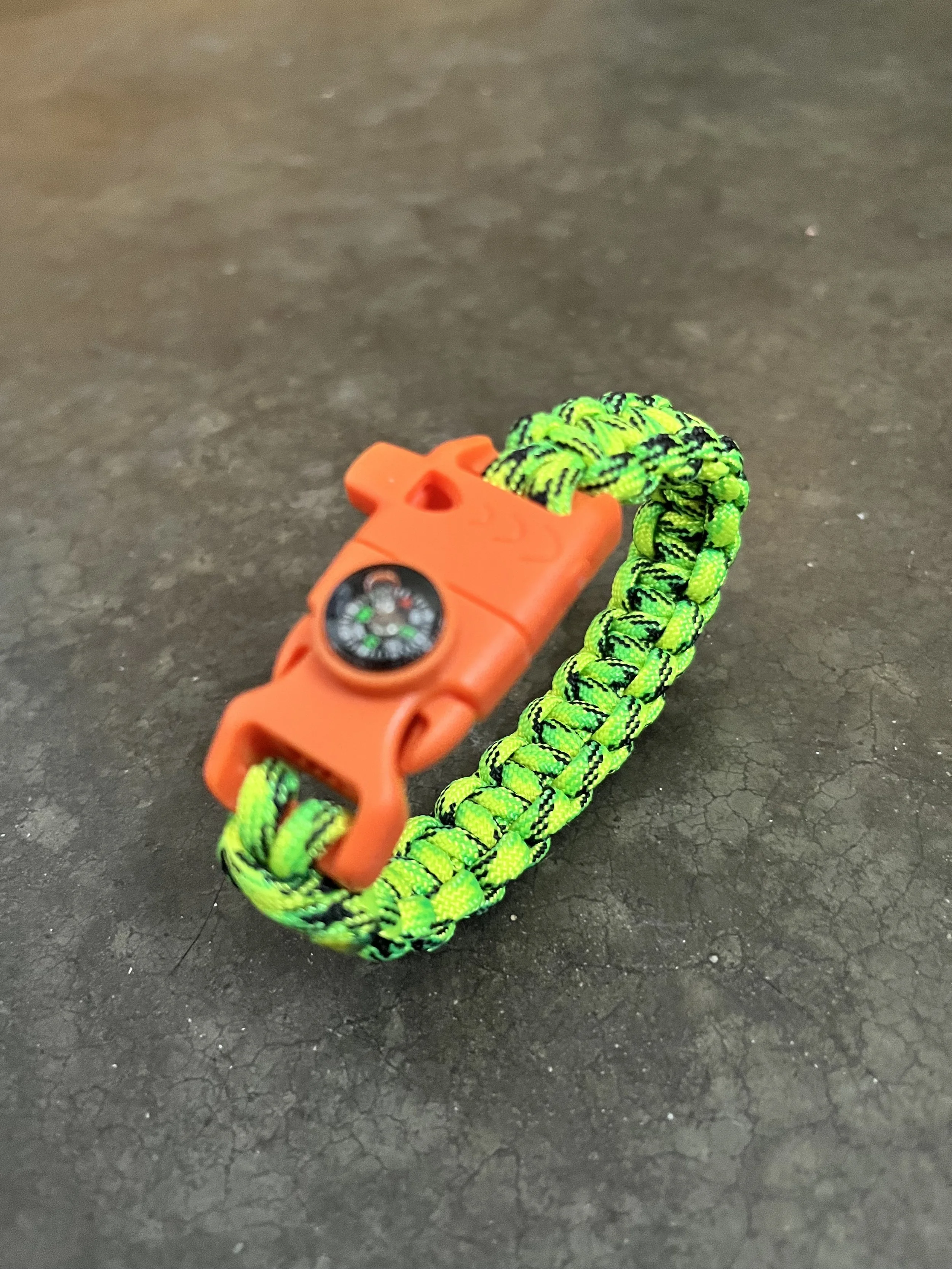 A close-up of a green and black braided paracord bracelet with an orange compass/whistle.
