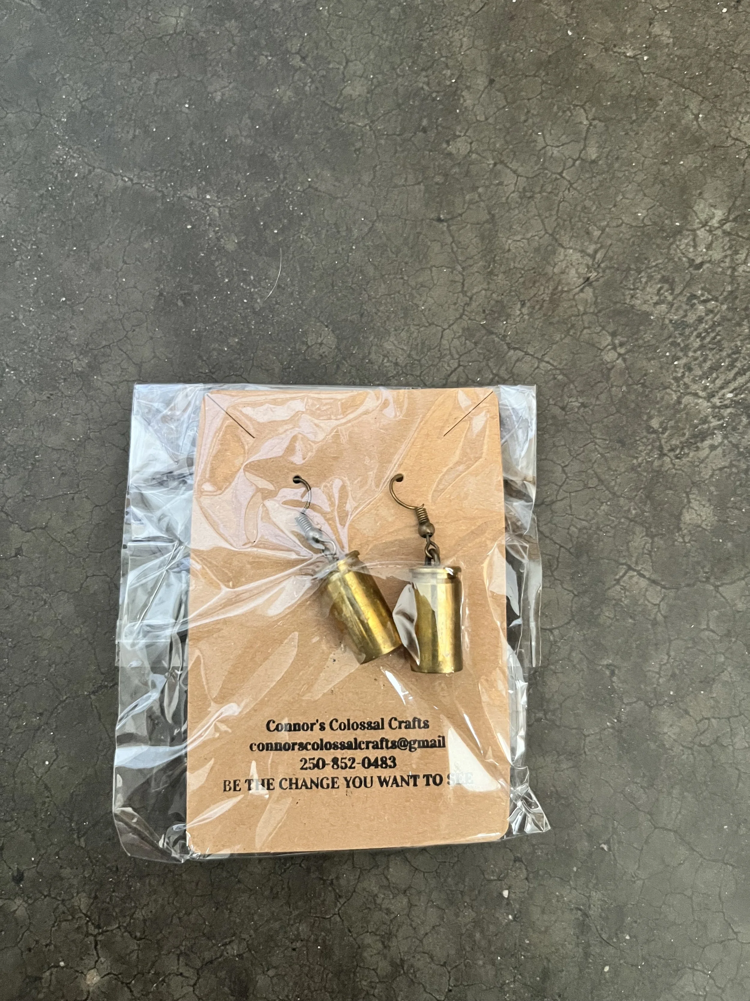 A clear plastic bag containing two gun casing earrings placed on a piece of cardboard with contact information for Connor’s Colossal Crafts on a cracked concrete surface.