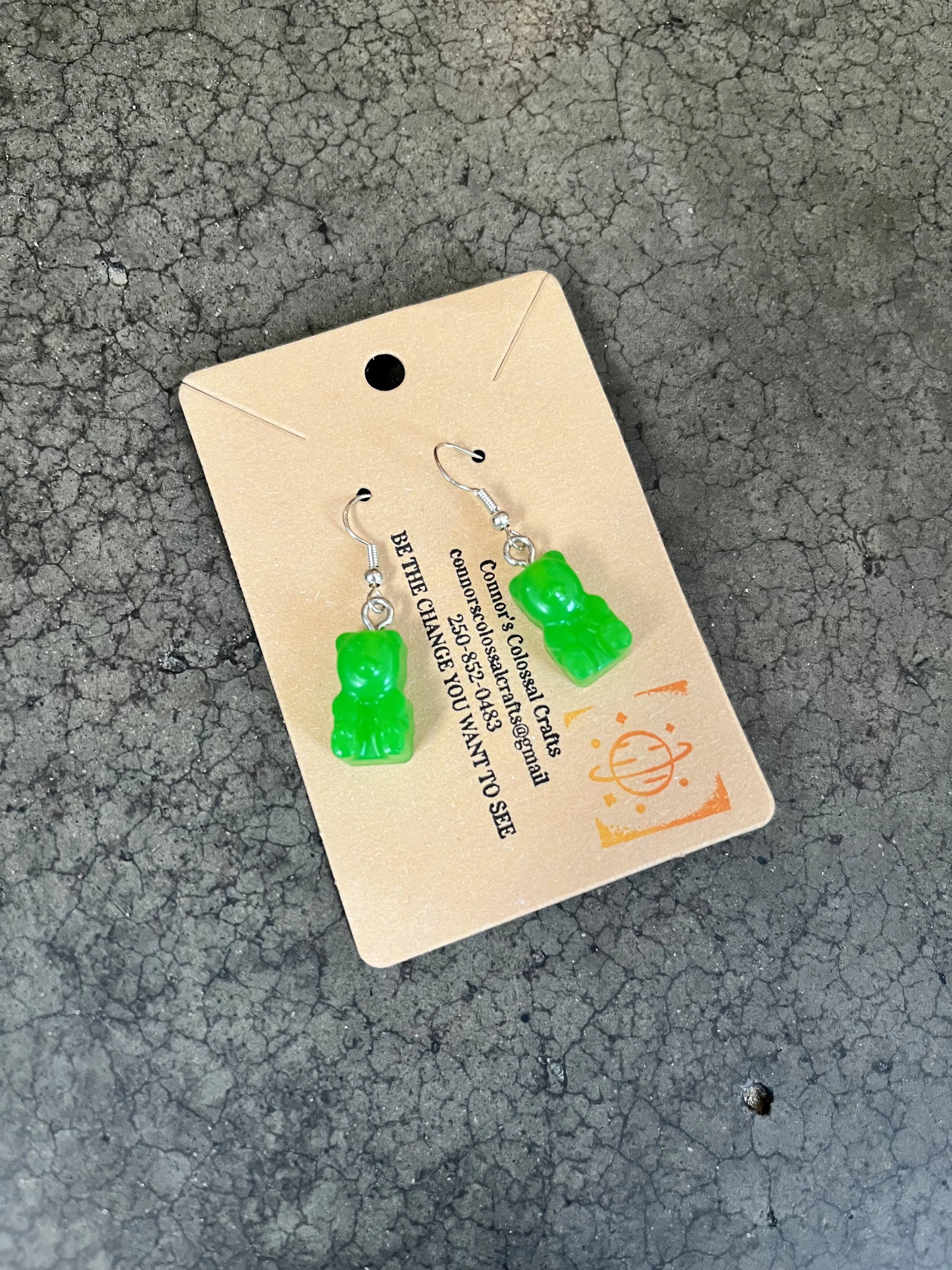 Green gummy bear earrings on a beige card, placed on a rough asphalt surface.