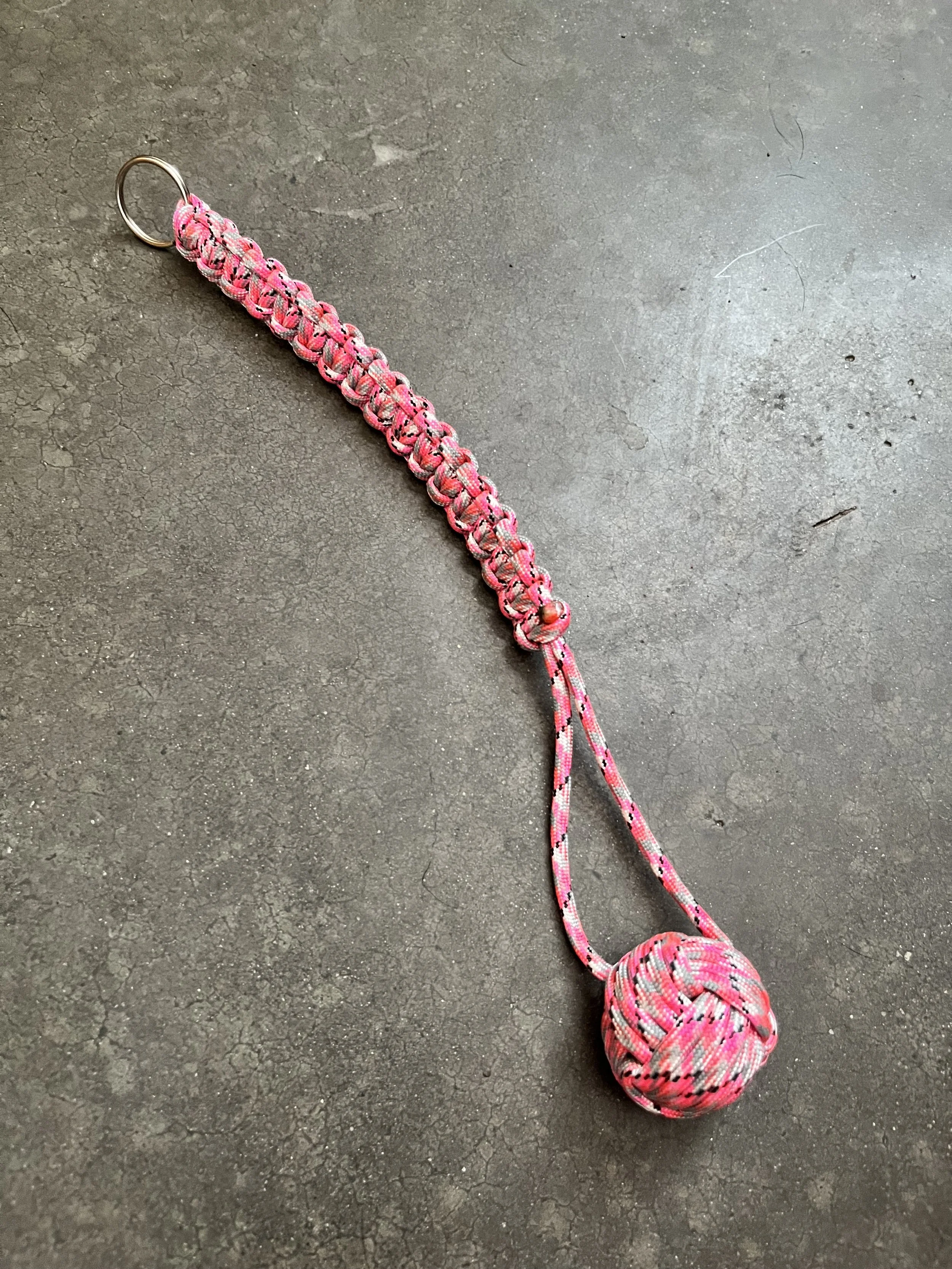 A pink and gray paracord dog leash with a knot at the end, lying on a dark textured floor.