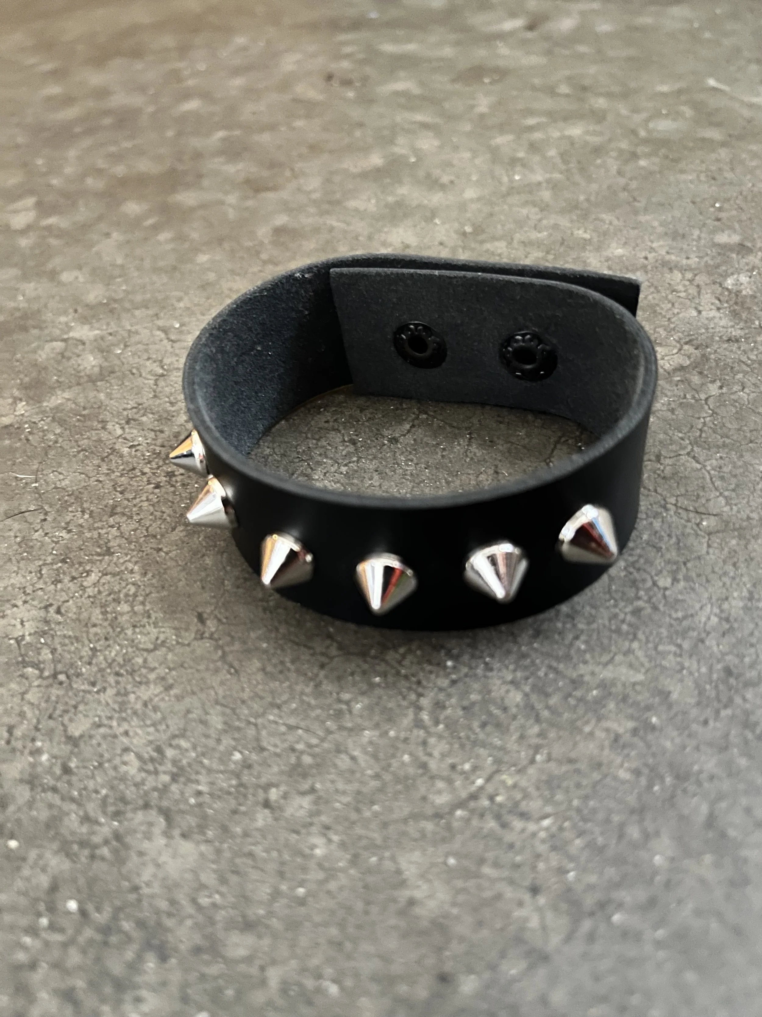 A handmade black fake leather bracelet with silver conical spikes around it, placed on a textured gray surface.