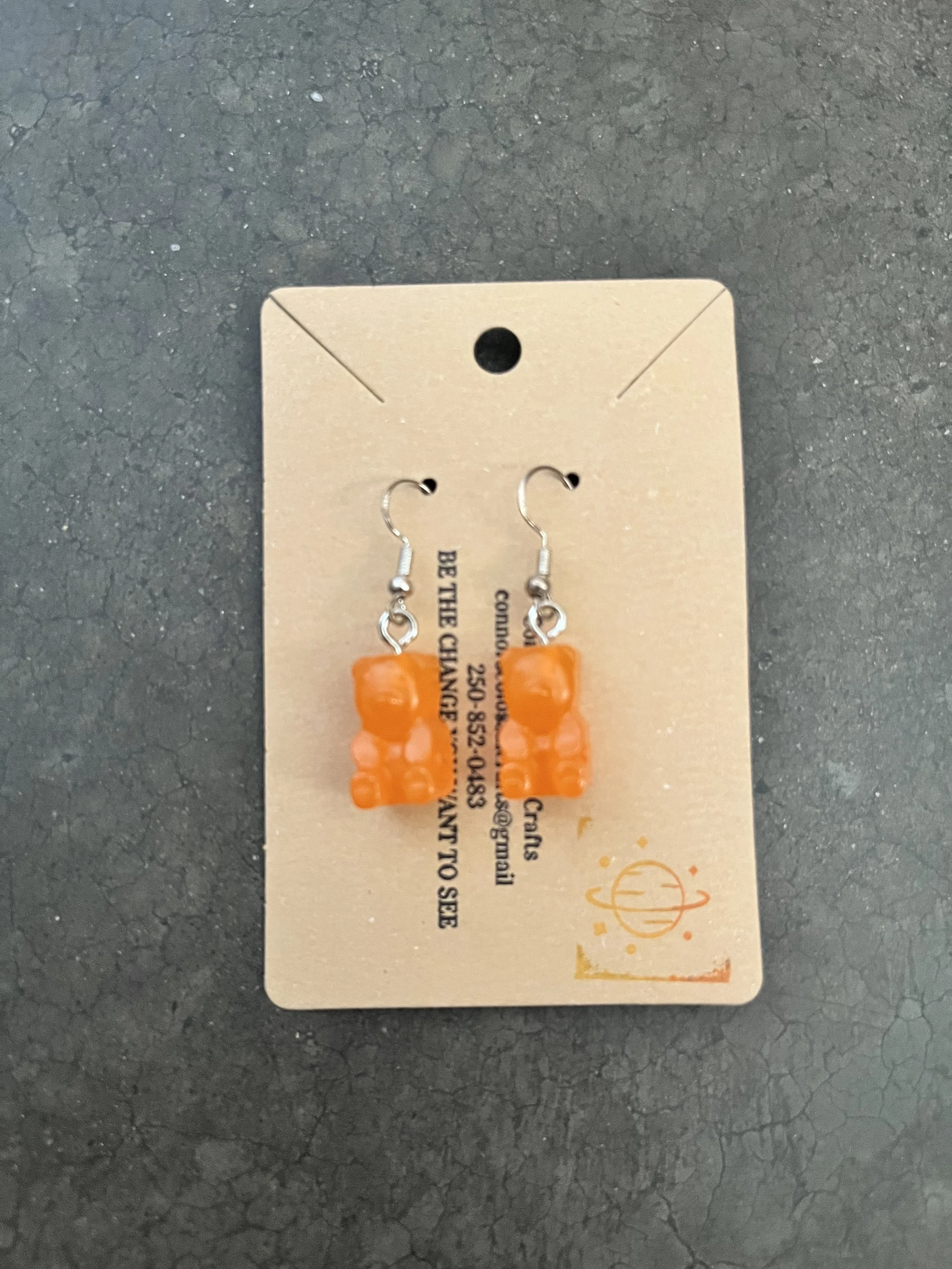A handmade pair of orange gummy bear earrings on a beige cardboard backing.