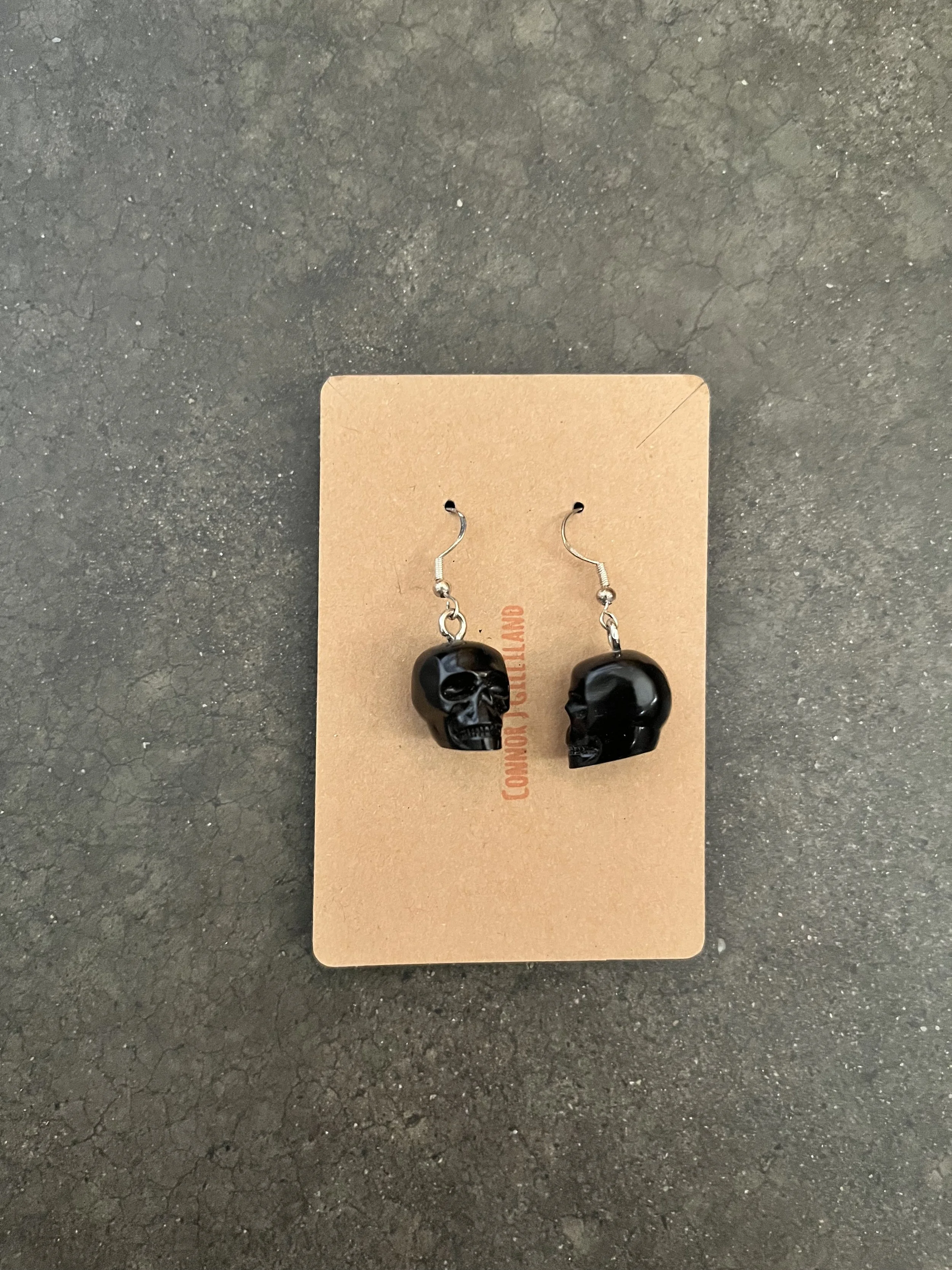 Black skull earrings displayed on a brown cardboard backing on a dark textured surface.