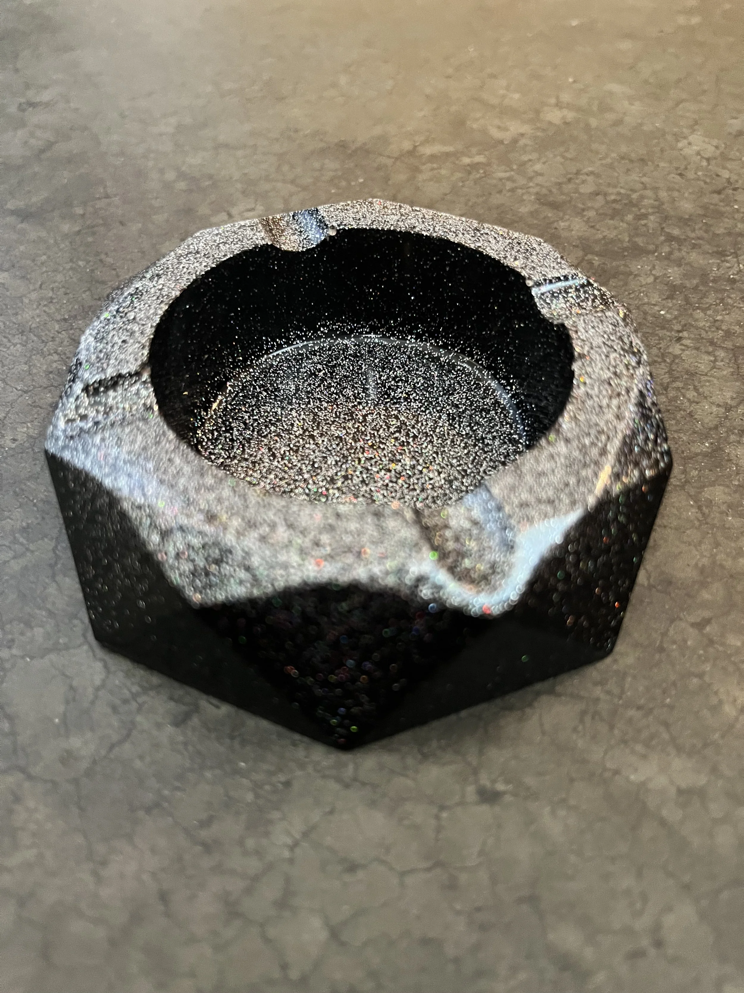 Close-up of a black and silver glittery ash tray with a rounded opening, placed on a gray textured surface.