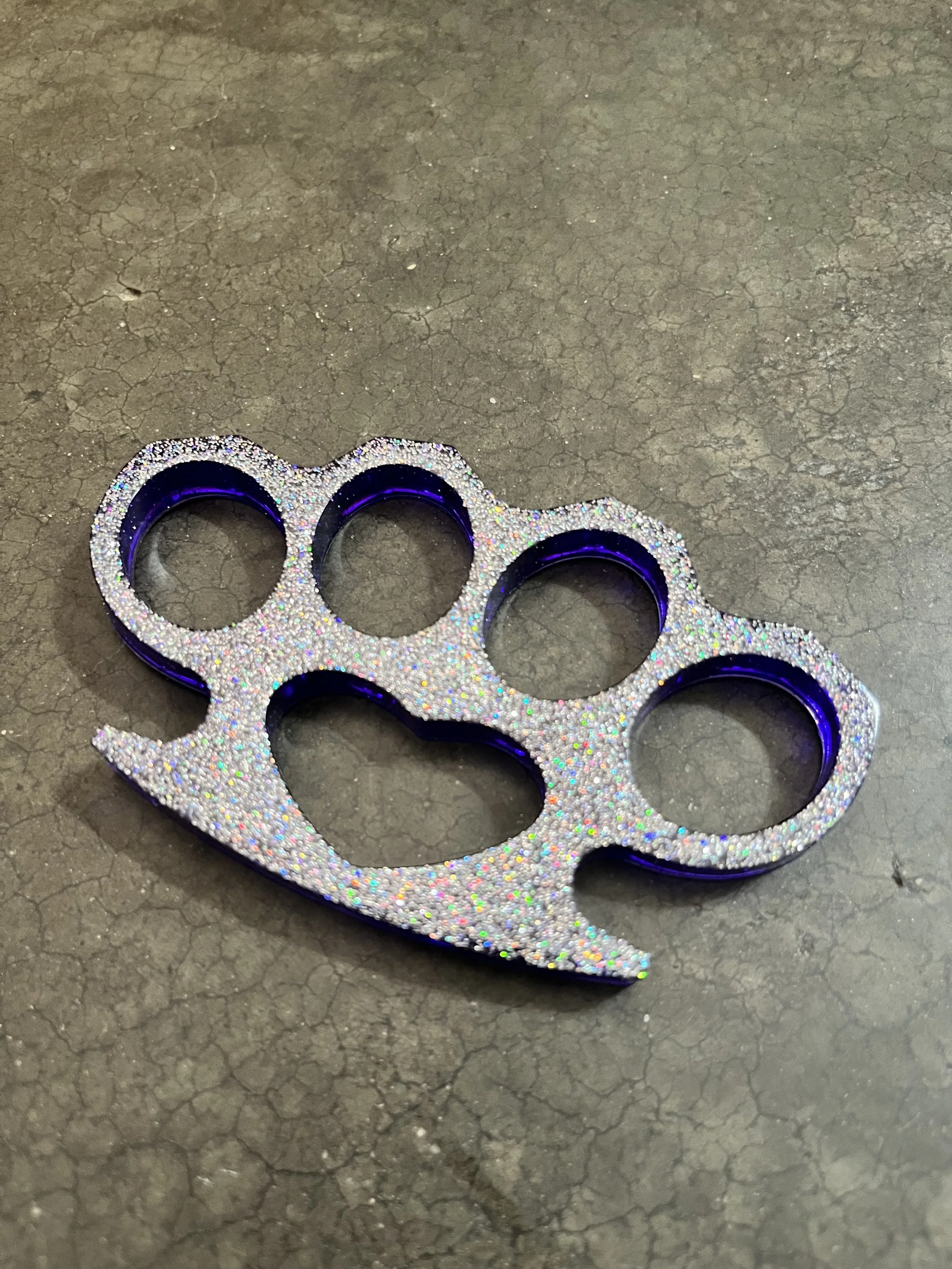 A metallic brass knuckle ring decorated with multicolored glitter, placed on a cracked gray surface.