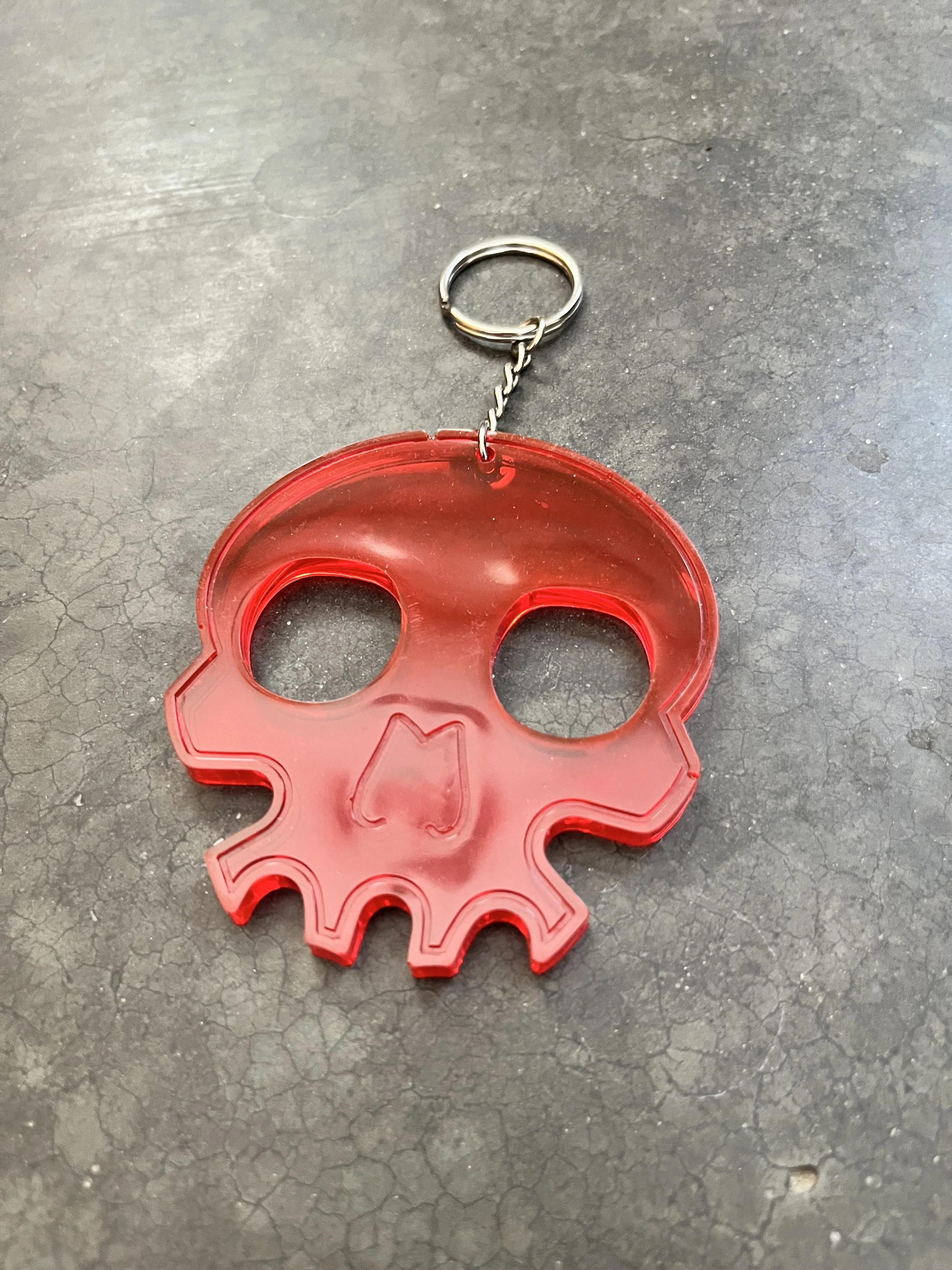 A red skull-shaped keychain with a small metal chain attached to a circular keyring on a dark, cracked surface.