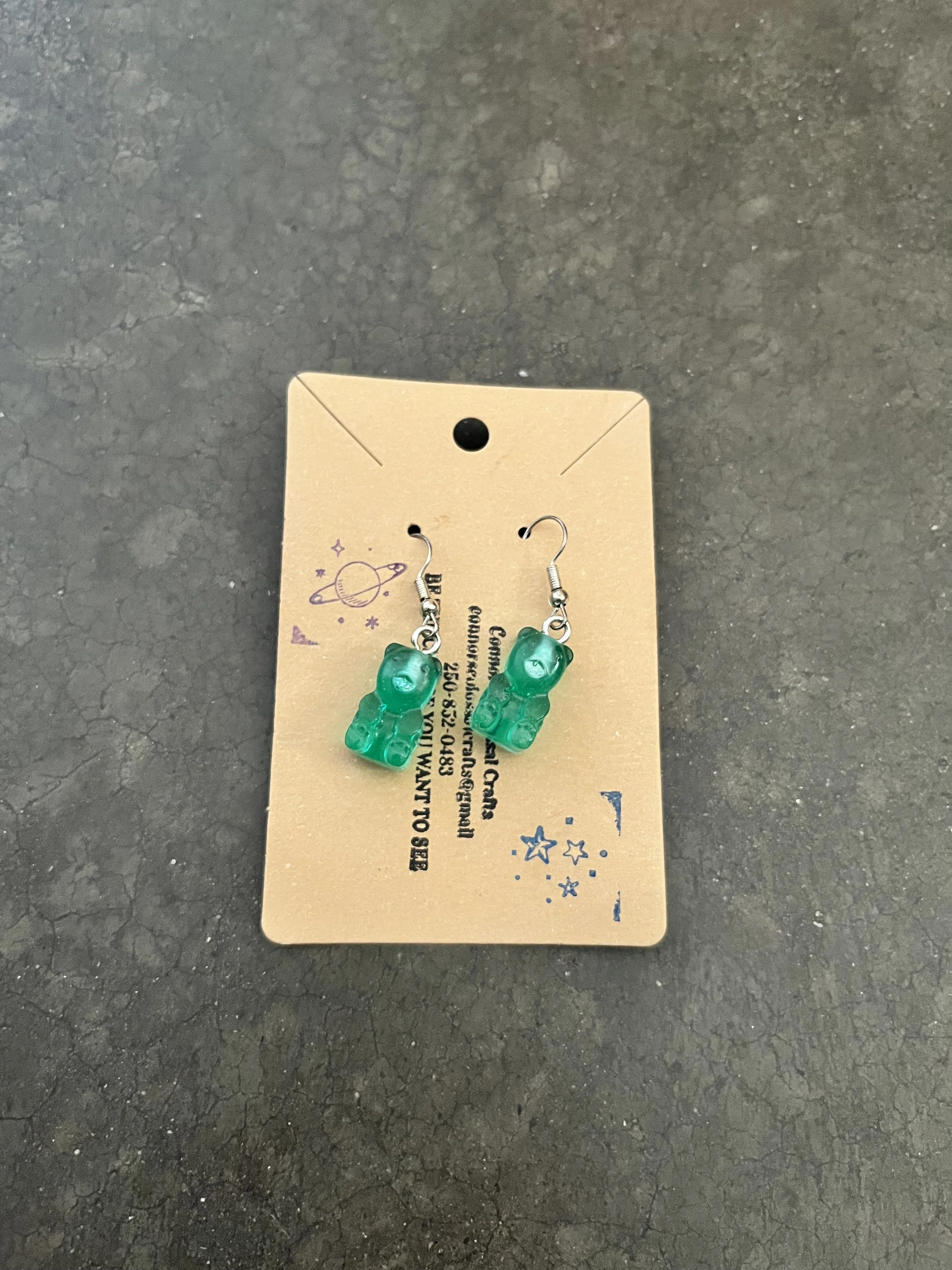 A handmade pair of green/teal gummy bear earrings on a cardboard backing.