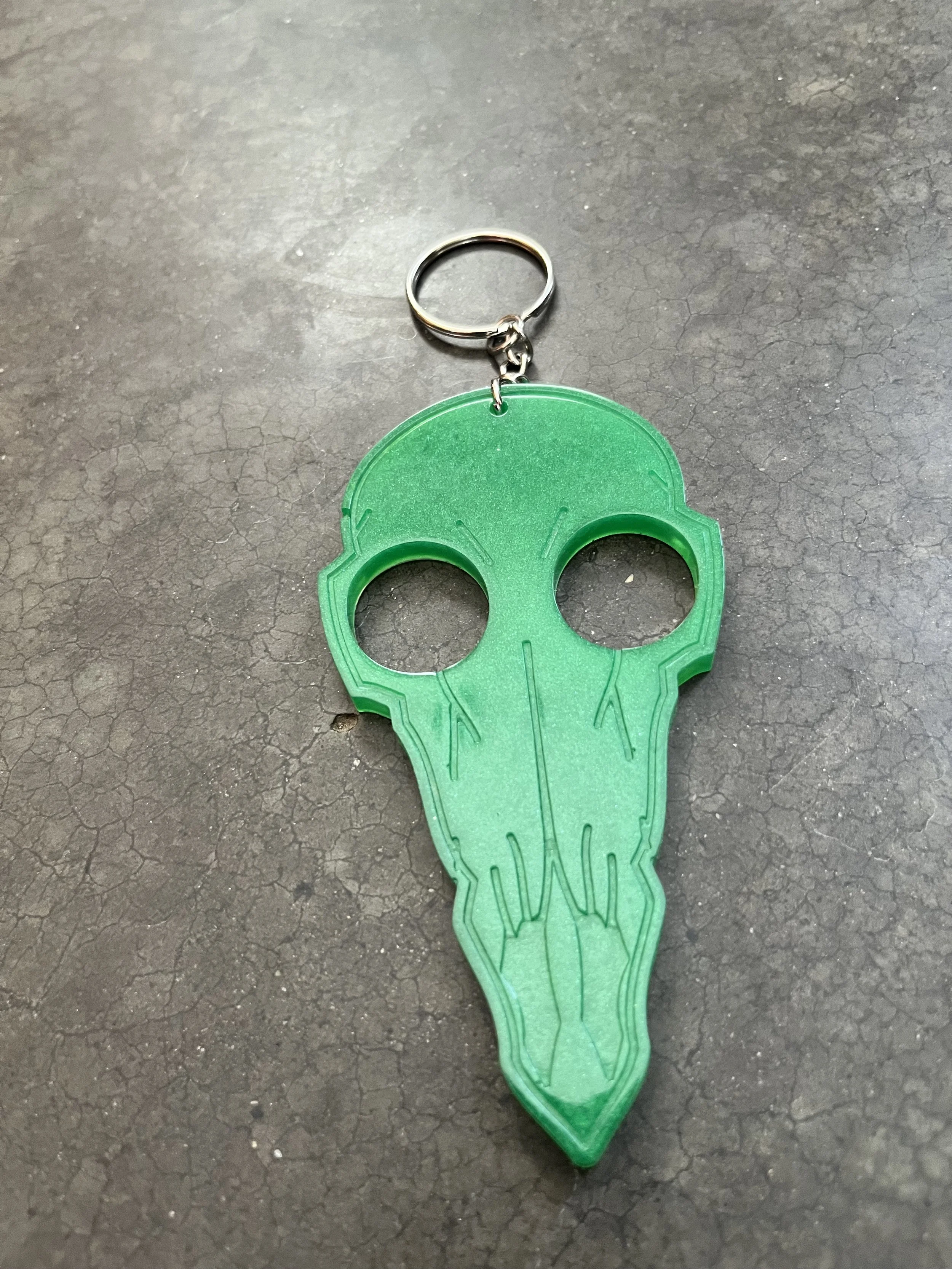 A green plastic keychain shaped like a skull with hollow eye sockets and elongated teeth outline, attached to a metal keyring, resting on a dark textured surface.