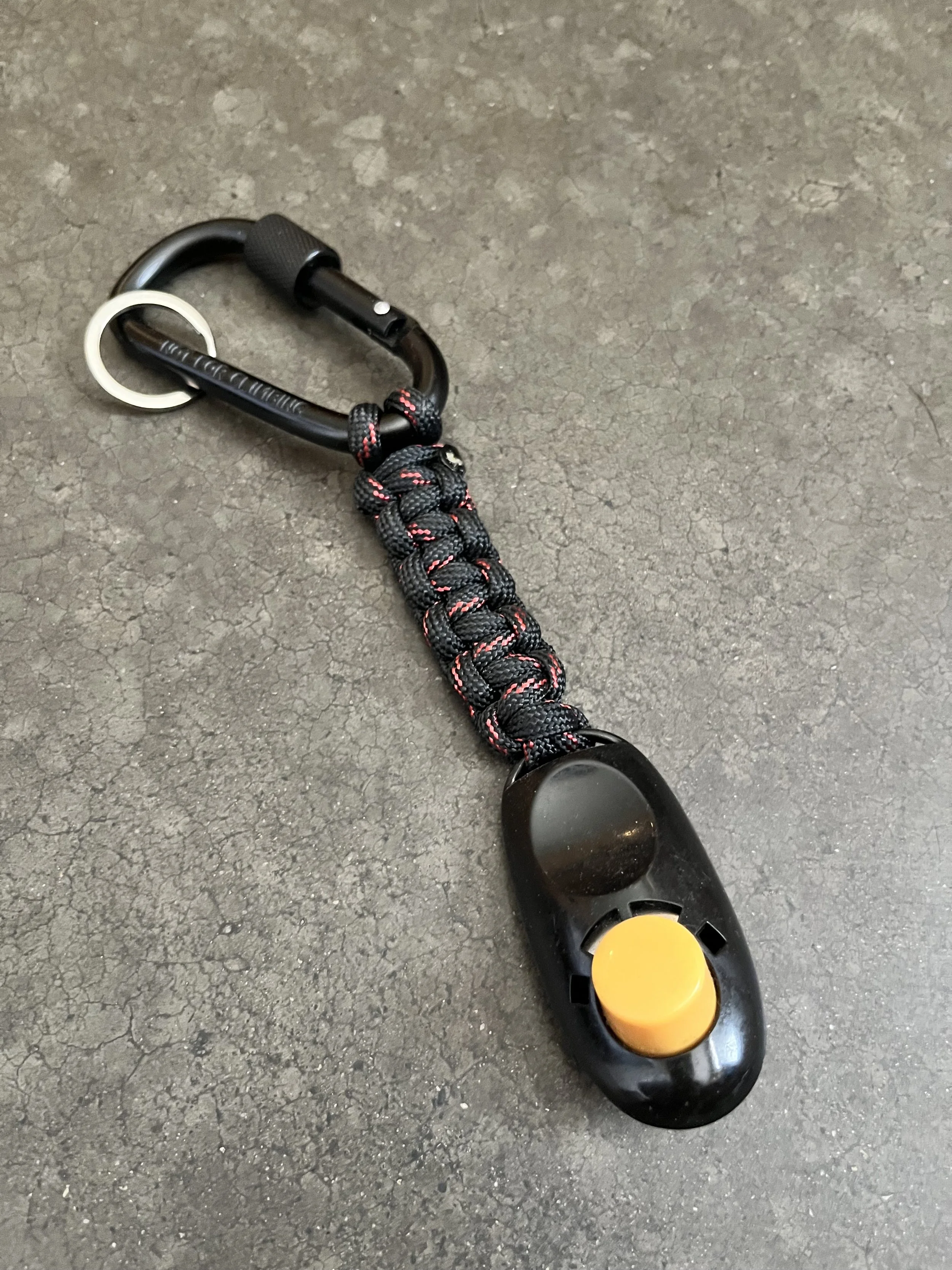 Close-up of a black plastic weight plate with a yellow center, attached to a black and red rope with a metal carabiner, on a rough concrete floor.