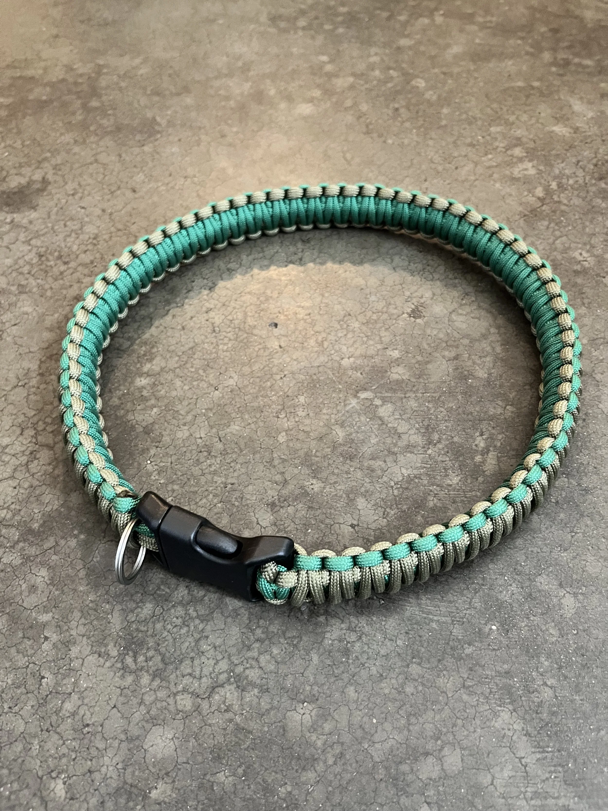 A green and gray paracord dog collar with a black plastic clip and metal ring on a cracked concrete surface.