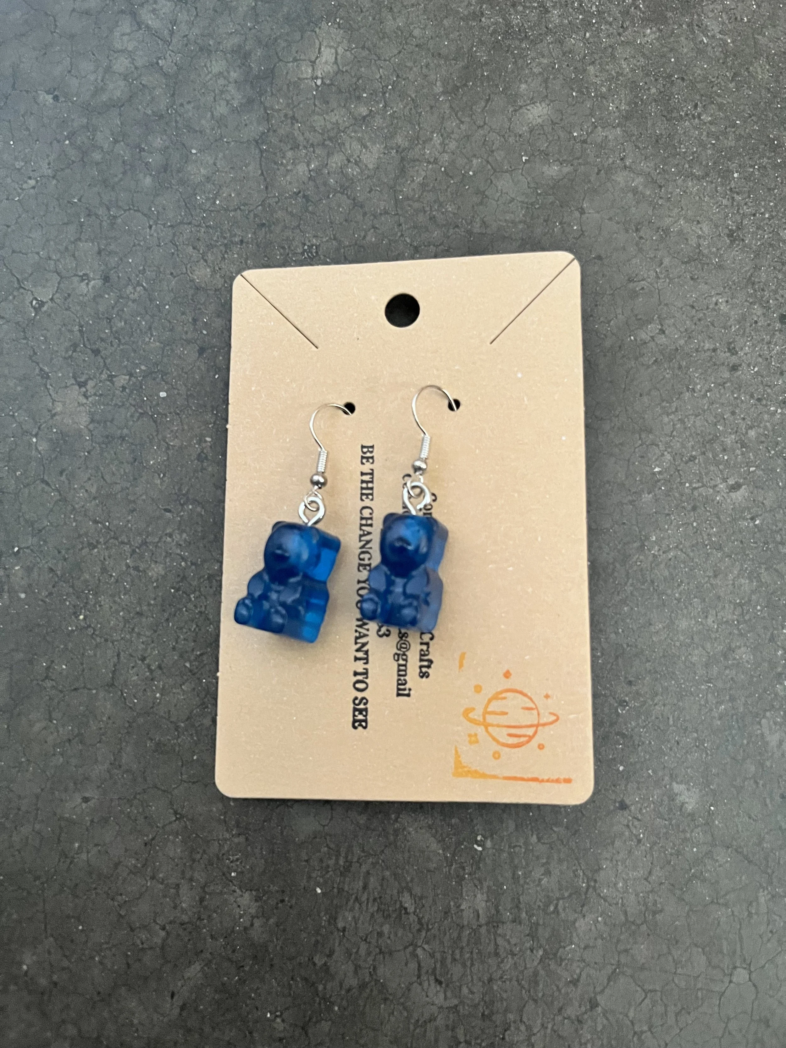Blue gummy bear earrings hanging from a cardboard backing with black text and an orange logo, on a dark, cracked surface.