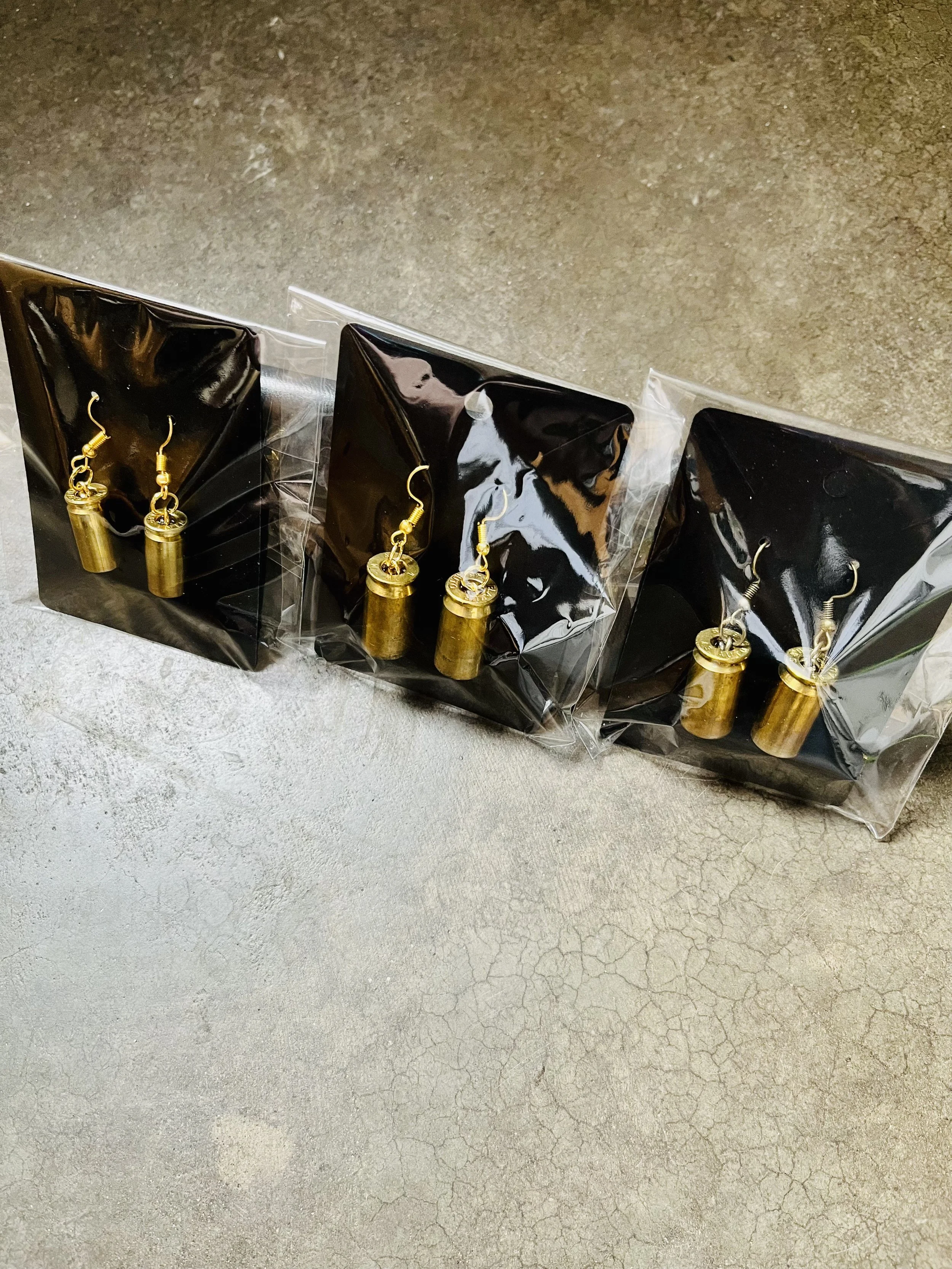A display of gun casing earrings on black cardboard backing, in clear plastic packaging, on a cracked concrete floor.