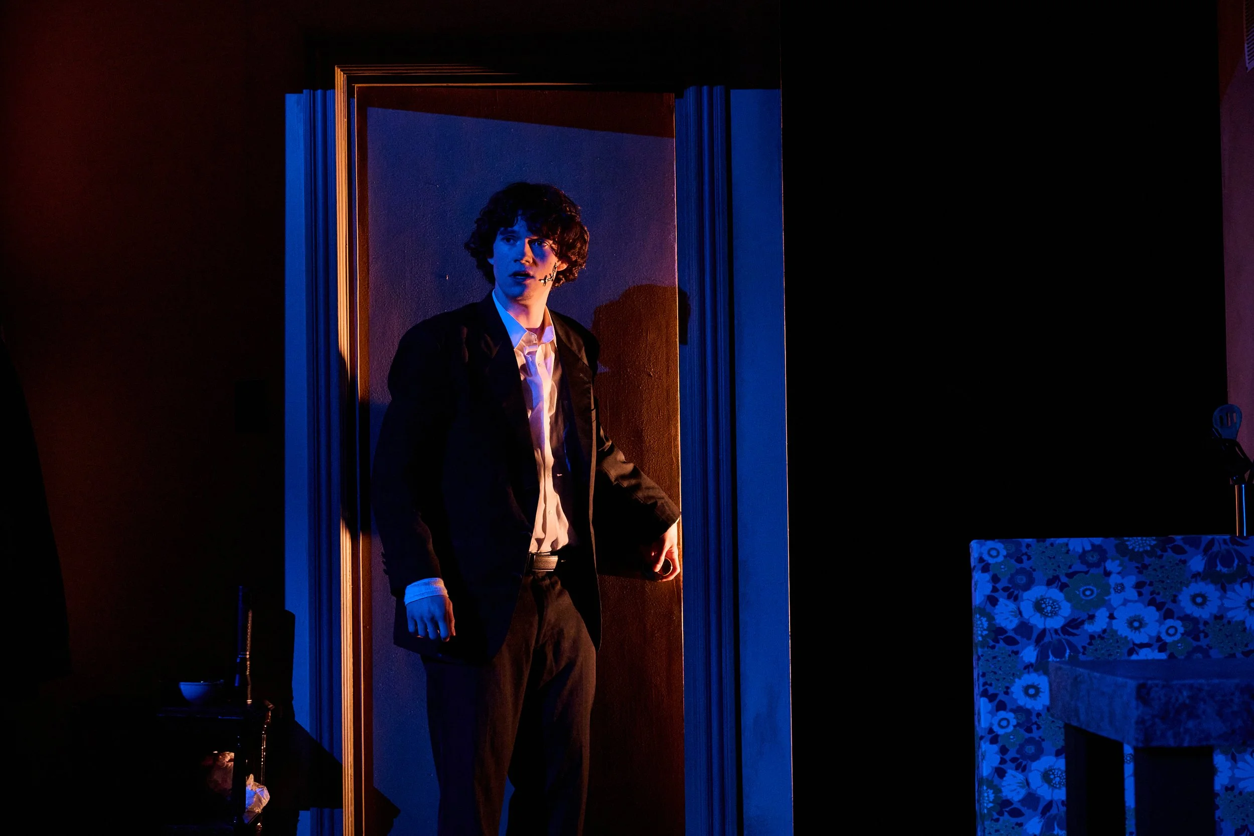 A young man with curly hair stands in a dimly lit room, wearing a dark jacket over a white shirt, looking emotional with his mouth slightly open and one hand holding a cellphone.