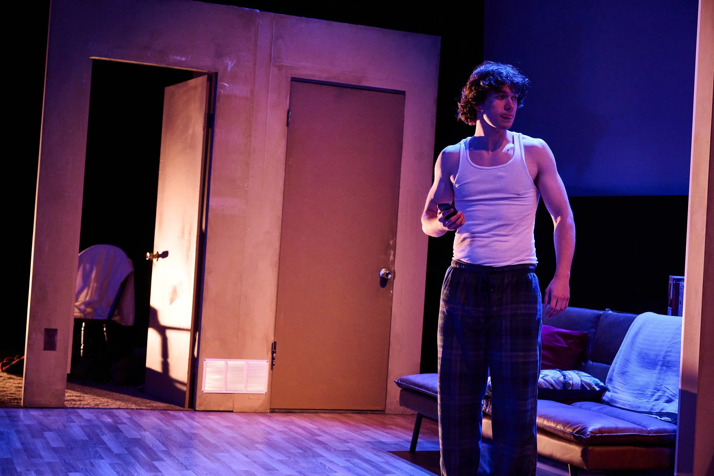 A young man with curly hair wearing a white tank top and plaid pajama pants standing in a dimly lit room, holding a remote control or phone, looking to his right with his tongue slightly out, in front of a wooden room divider and a couch with pillows