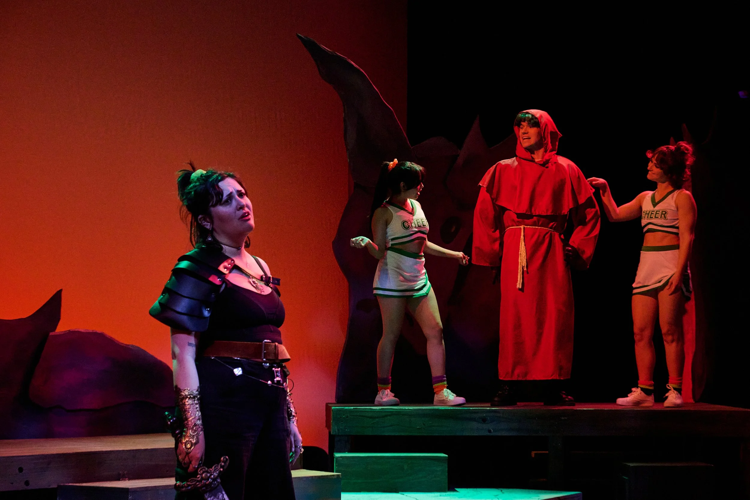 Scene from a theatrical performance with four young women and one young man on stage. The women are dressed in cheerleading outfits, and the man is dressed in a red hooded cloak. The background has warm lighting and artistic set pieces.
