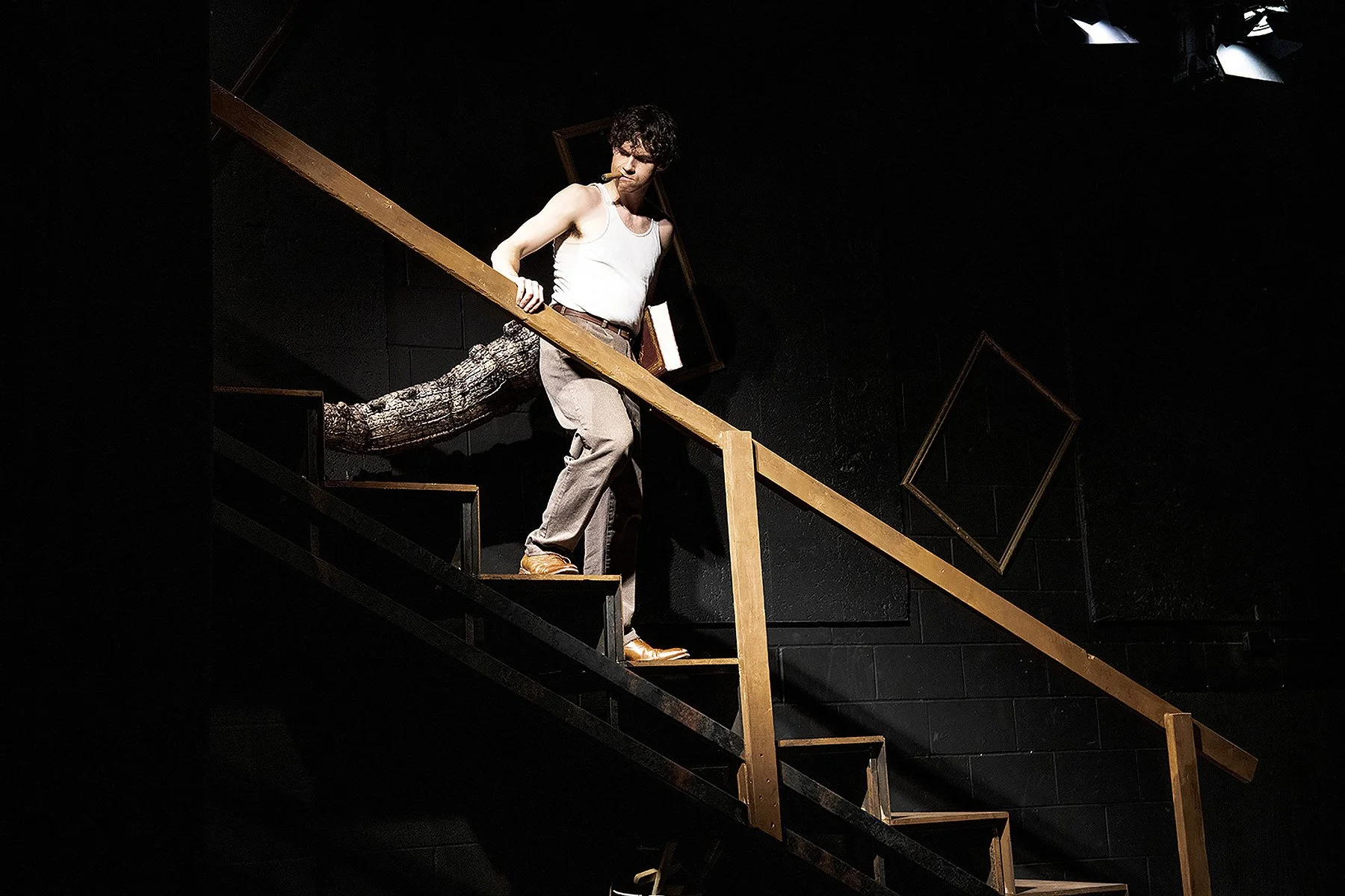 A man with dark, curly hair wearing a white tank top, beige pants, brown shoes, and a large tail, standing on a staircase with a wooden handrail. He is holding the handrail with one hand, has a cigar in his mouth, and appears to be looking downward,.