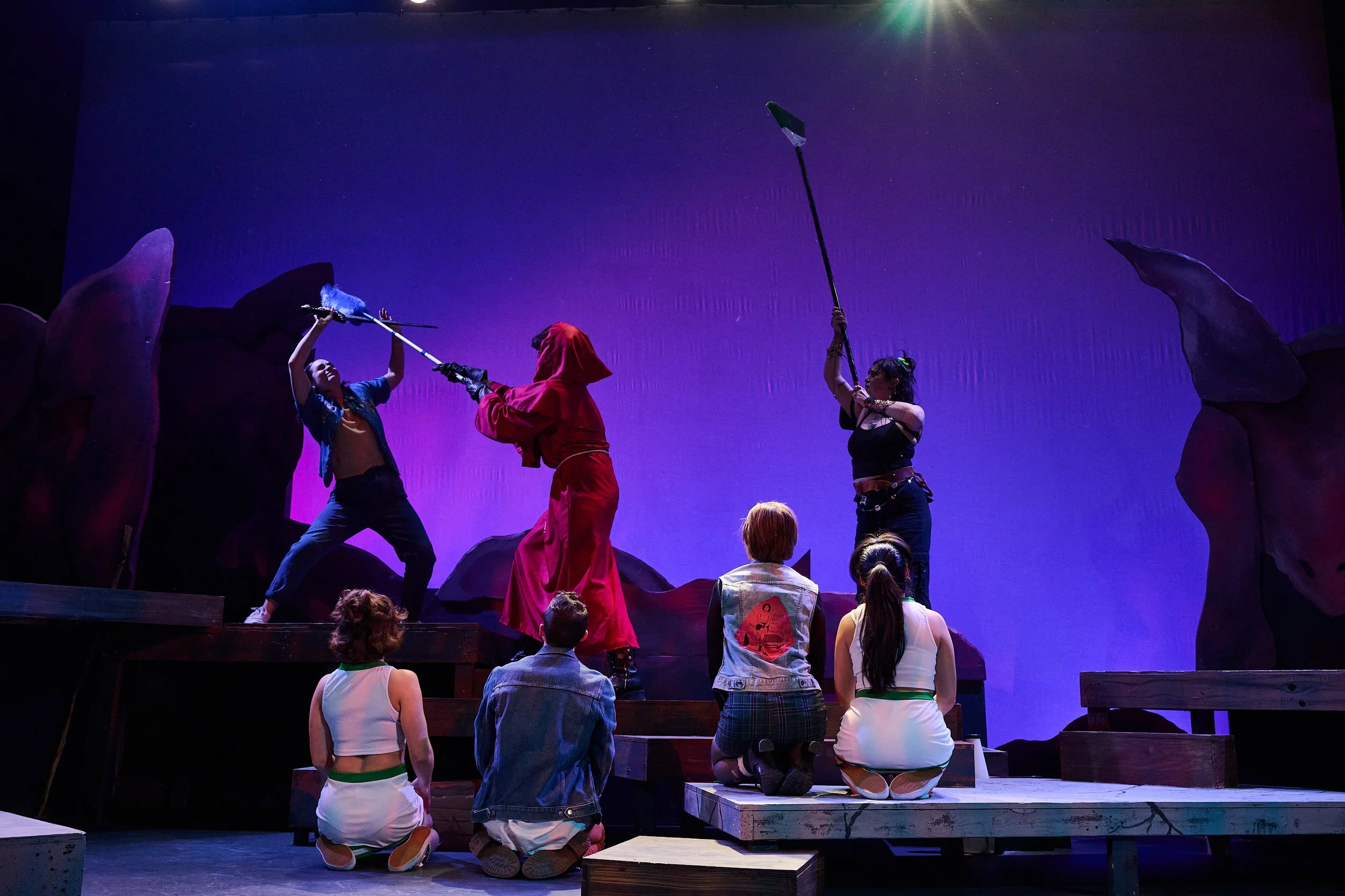 Stage performance of three actors dressed in costume, with animated background and audience members watching. One actor is in red with a cape, another is in black, and the third is in blue, holding a spear or staff. Audience members are kneeling or s