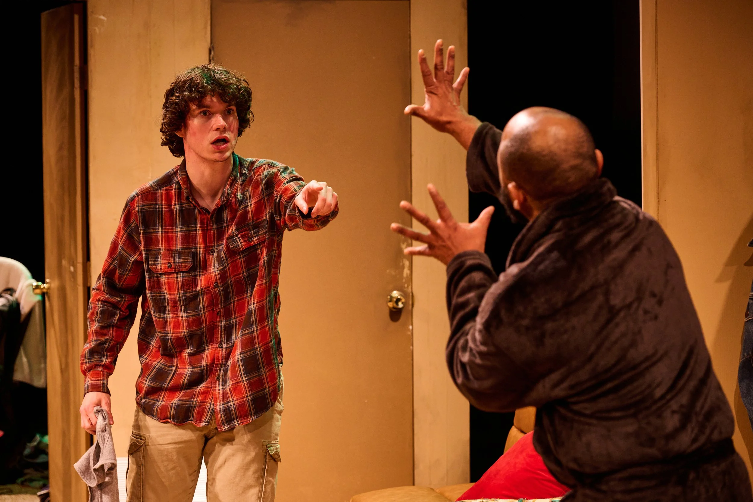 A young man with curly hair wearing a red plaid shirt and khaki pants is speaking or explaining something to an older man with a shaved head, who is raising his hand and wearing a dark brown jacket.
