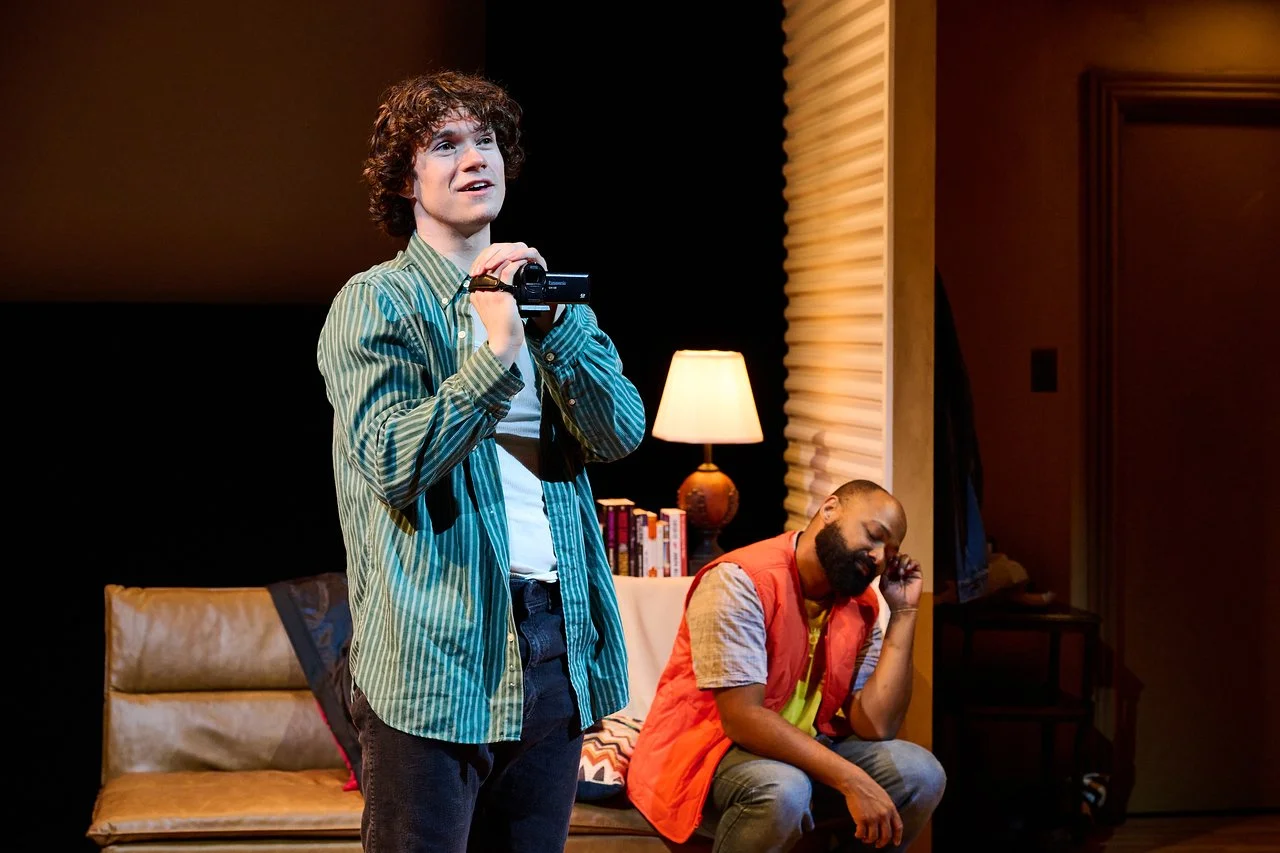 A young man with curly hair and striped shirt holding a camcorder, looking upward. In the background, a bearded man with a shaved head in an orange vest sitting on a sofa, resting his head on his hand, appearing contemplative. The setting is a cozy r