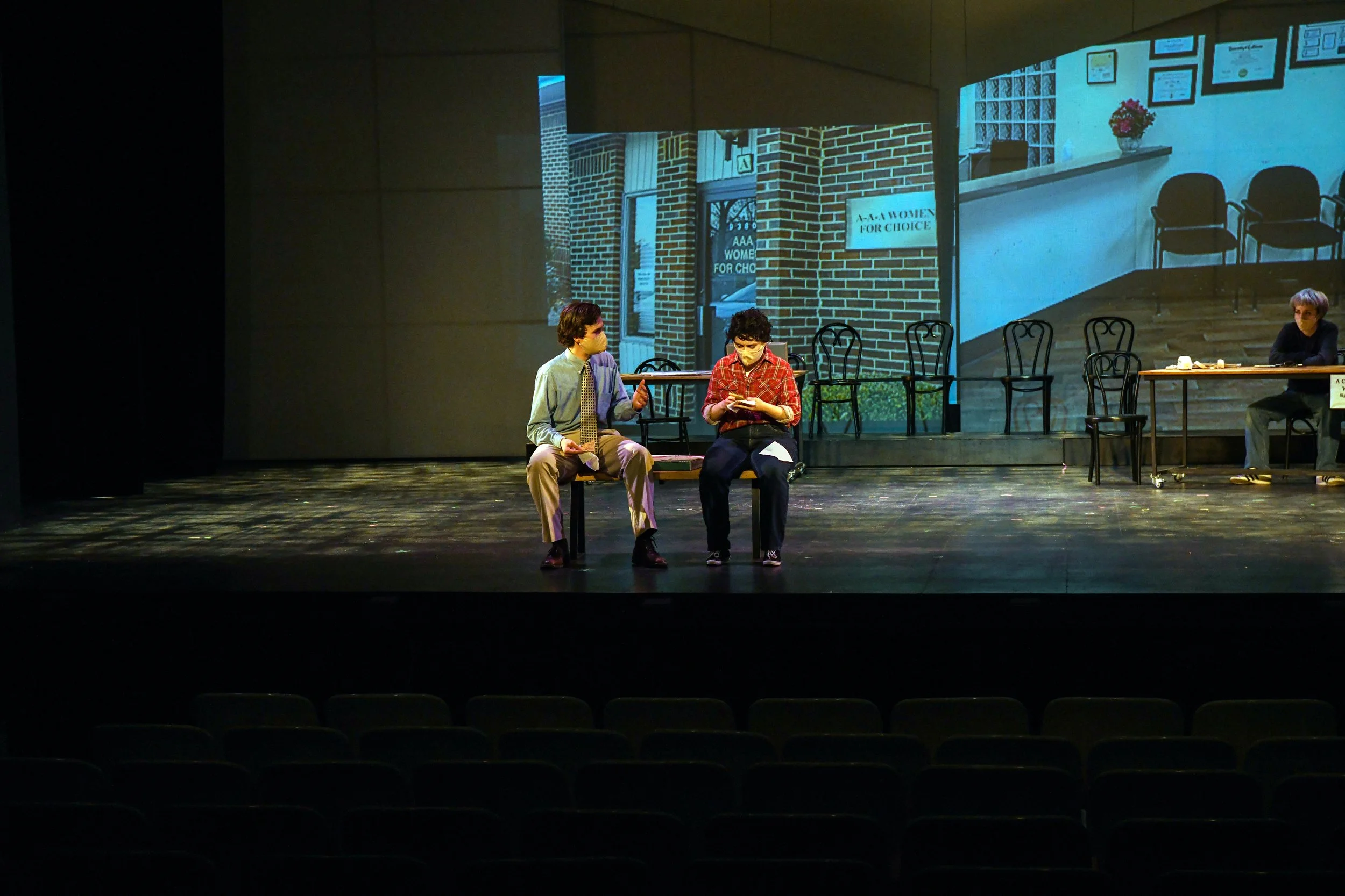 A stage with two actors sitting on chairs, engaging in conversation, with a large backdrop projecting an outdoor scene of a brick building with a sign that reads "AAA Woman for Choice." In the background, a woman sits alone at a table on the stage, s