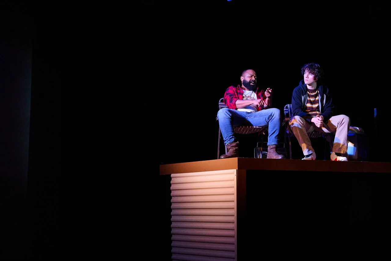 Two men sitting on chairs on a stage, engaged in a conversation, with a dark background and spotlight.