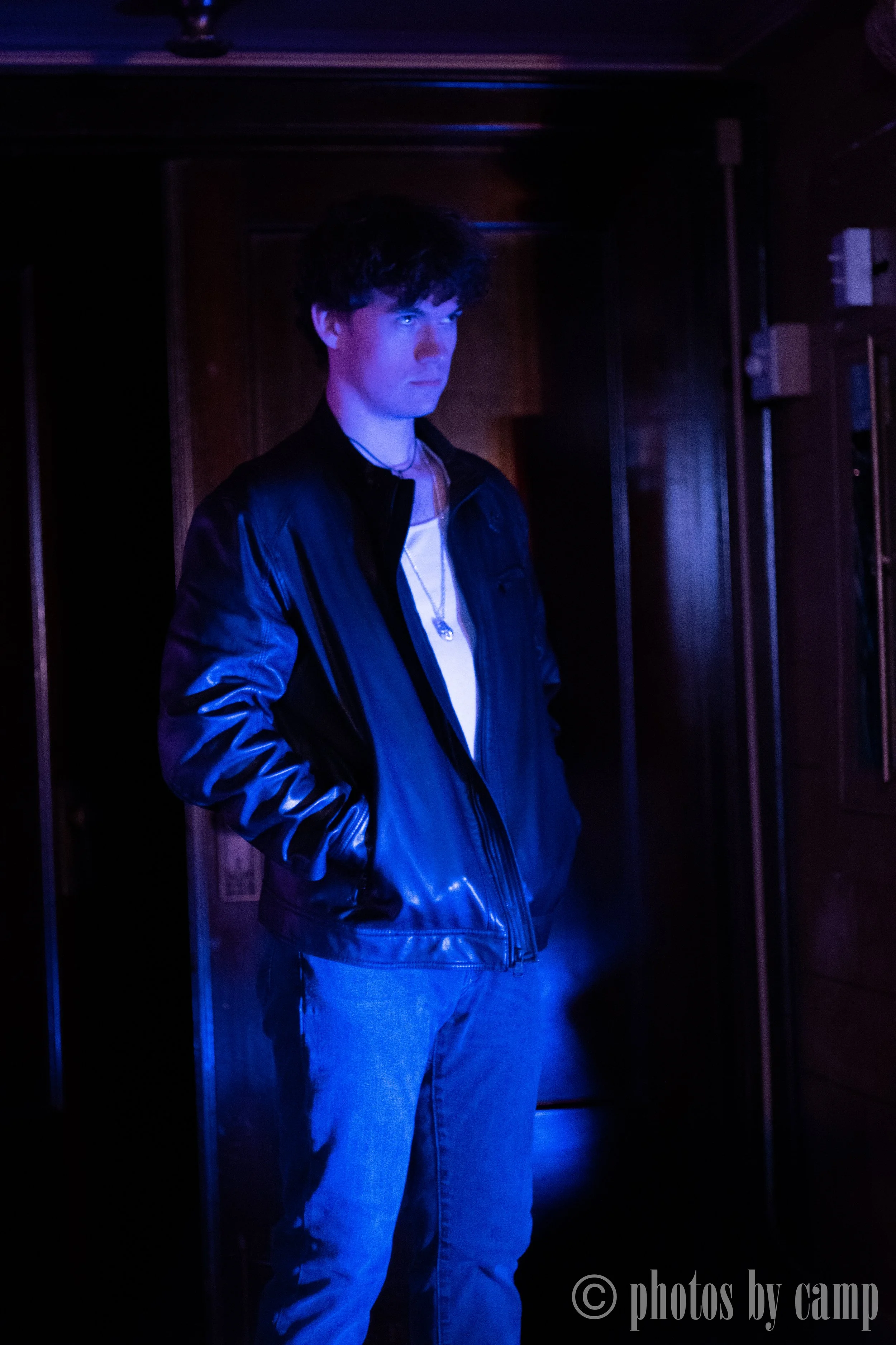 Teenage boy with dark curly hair wearing a black leather jacket, white t-shirt, and blue jeans standing in a dimly lit room with purple lighting.