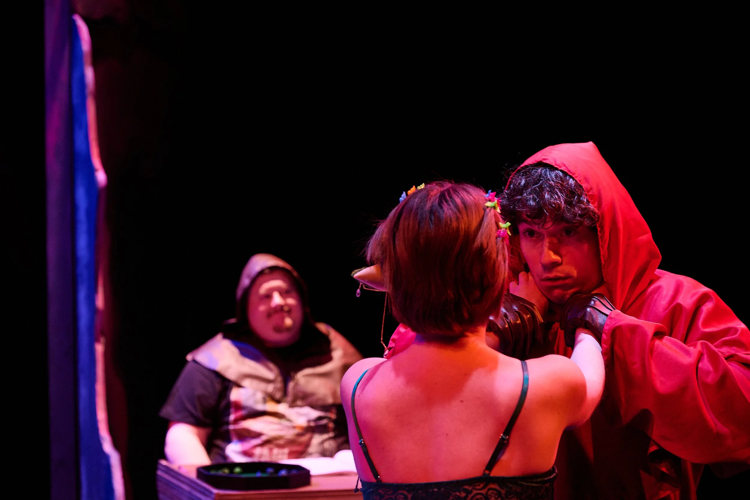 A group of performers on stage, with one person in a red hoodie leaning in close, possibly preparing for a theatrical scene. The scene is dimly lit with colorful stage lighting, with a black background and a partially visible mirror or prop on the le