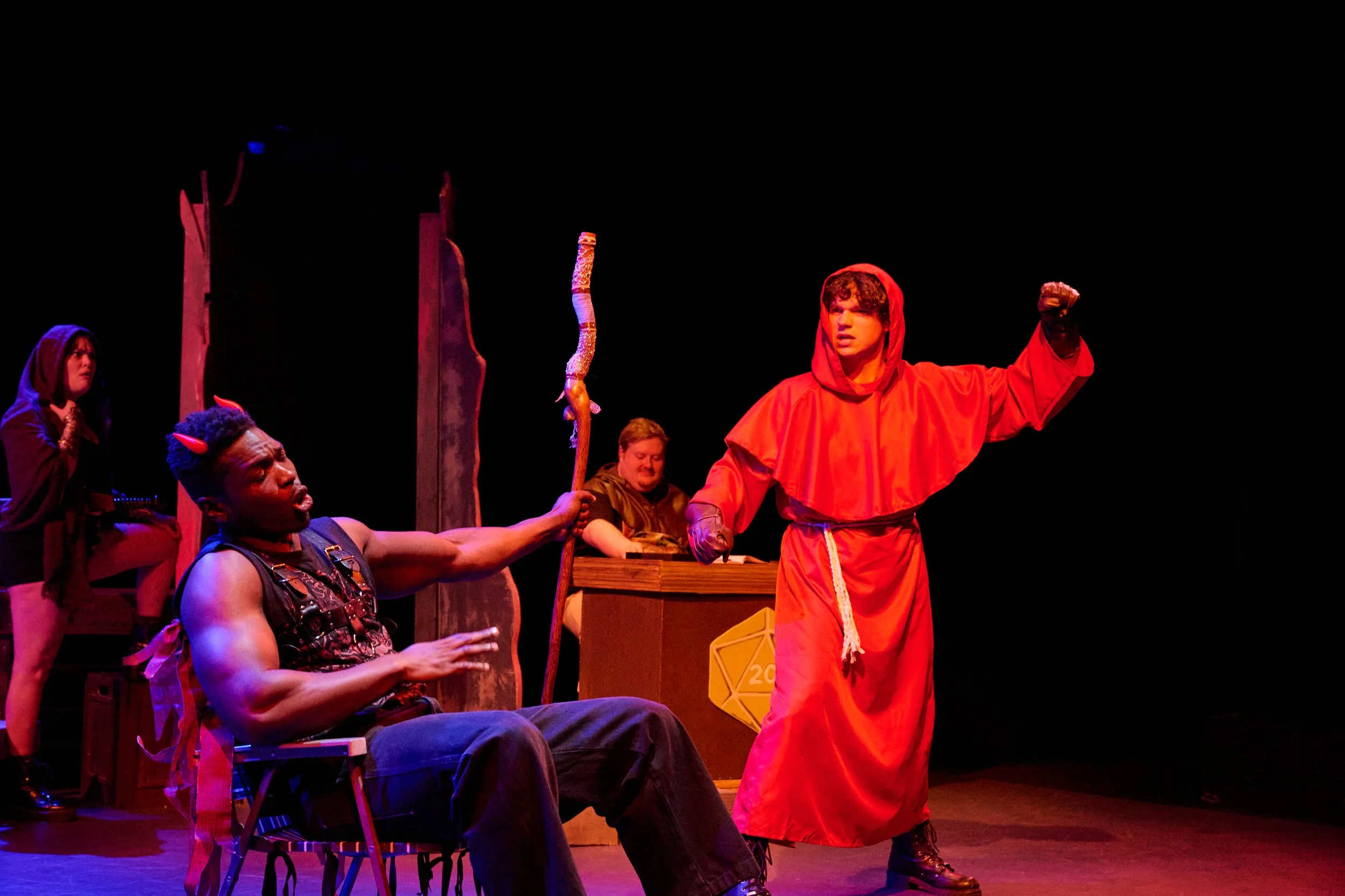 The image depicts a theatrical performance with five actors. One actor, dressed in a bright orange costume with a hood, is standing and raising their clenched fist. Other actors are seated or standing around, with some holding objects or props, and m