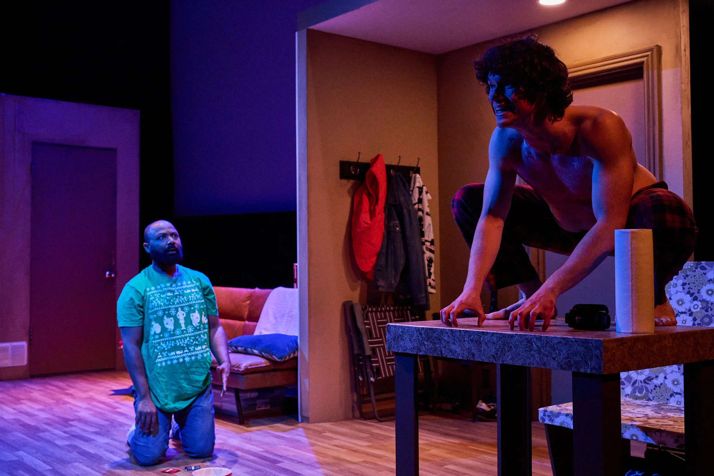 A woman with curly hair crouches on a table, smiling, with a man kneeling on the floor looking up at her. The scene is lit with colorful lighting in a cozy living room.