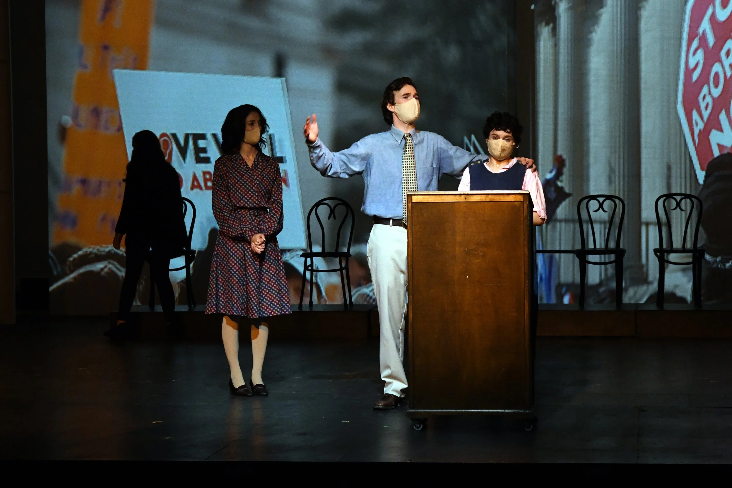 A stage with three people wearing masks, one man standing behind a wooden podium with his arm around a boy, and a woman standing to the side. A large screen in the background displays text and graphics. Chairs are arranged behind them.