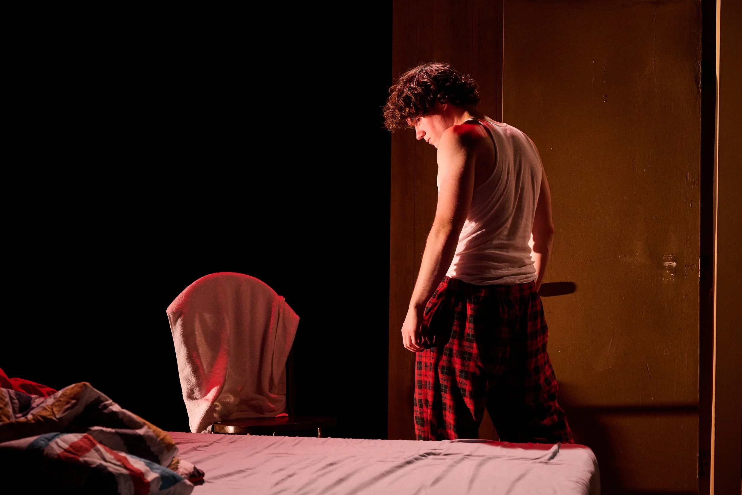 A person with curly hair standing in a dimly lit room wearing a tank top and plaid pajama pants, looking down. Another person with a hoodie over their head sitting on a bed with a colorful quilt. The bed has a white and red striped blanket. There is 