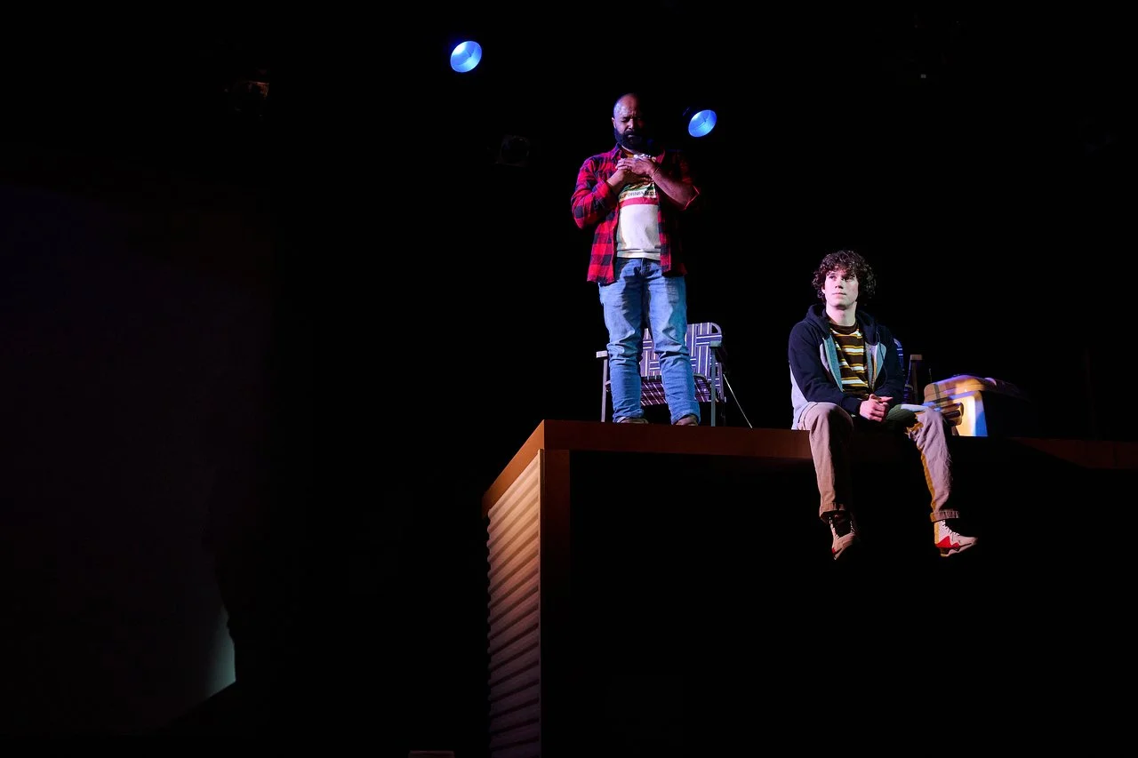 Two men on a stage, one standing and the other sitting, illuminated by stage lights, with dark background.