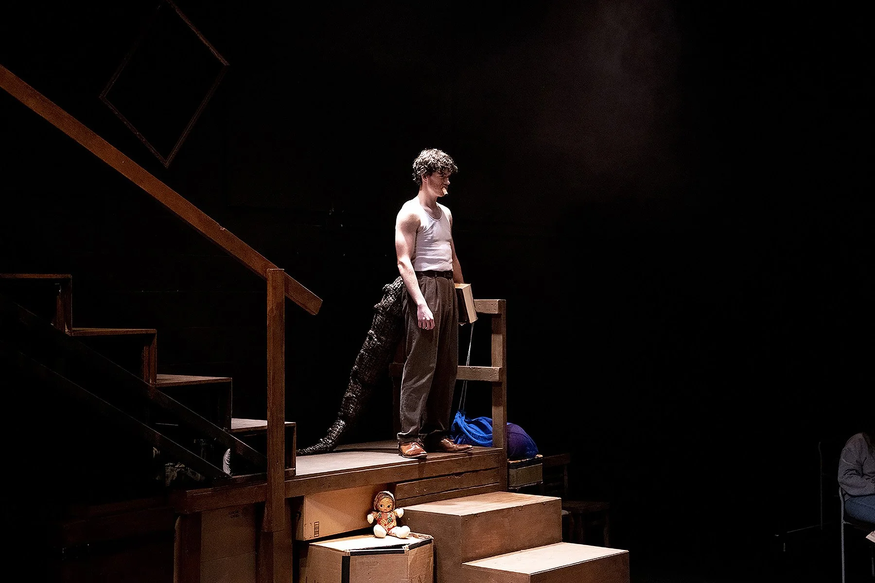 A young man stands on stage at the top of a wooden staircase, with part of a snake prop wrapped around his leg. He is wearing a white tank top, brown pants, and has curly hair. The stage is dark with a spotlight on him; there are various props and a 