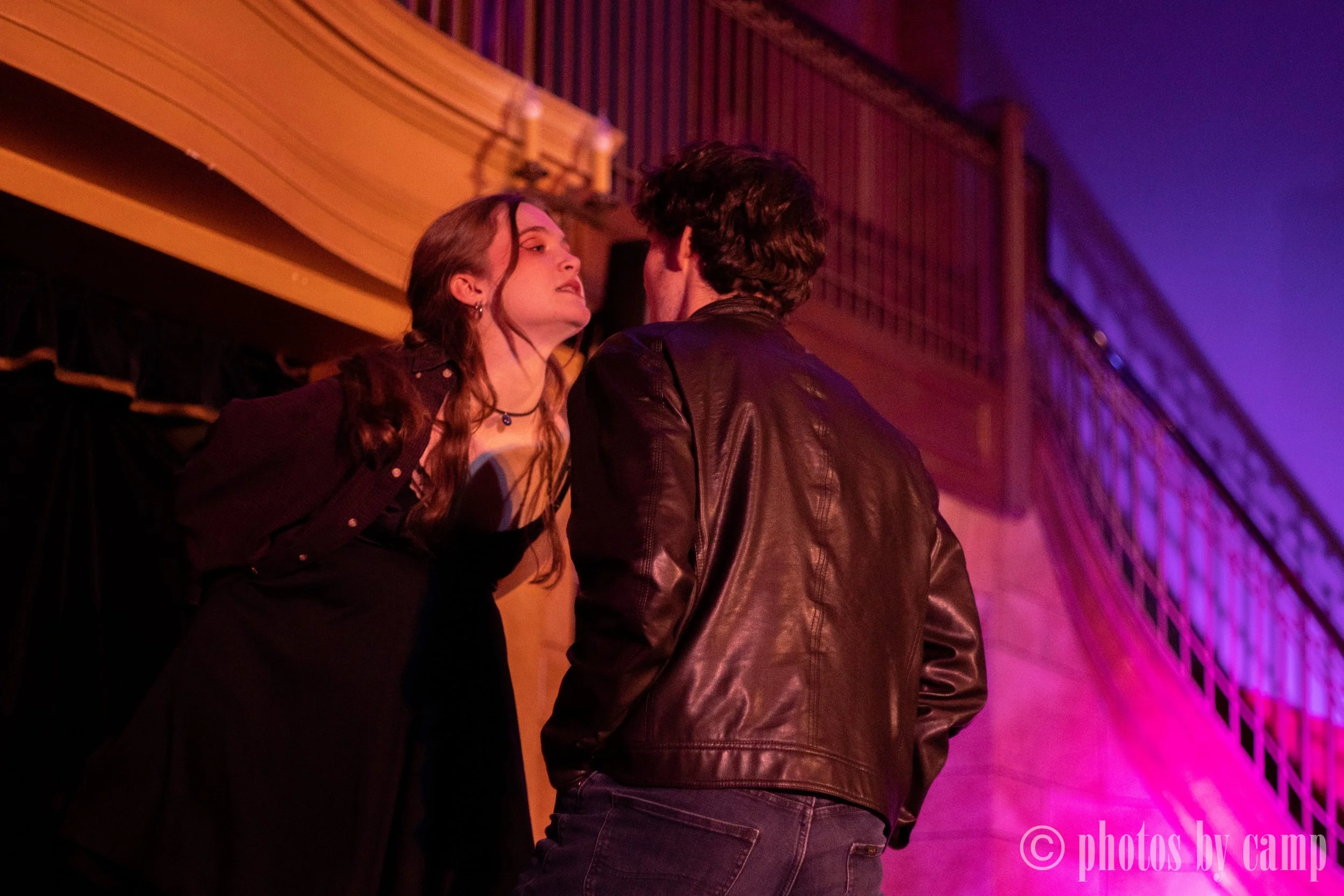 A young woman with long brown hair and a young man with dark curly hair stand close together indoors, engaging in a conversation with intense expressions, illuminated by colorful purple and orange lighting.