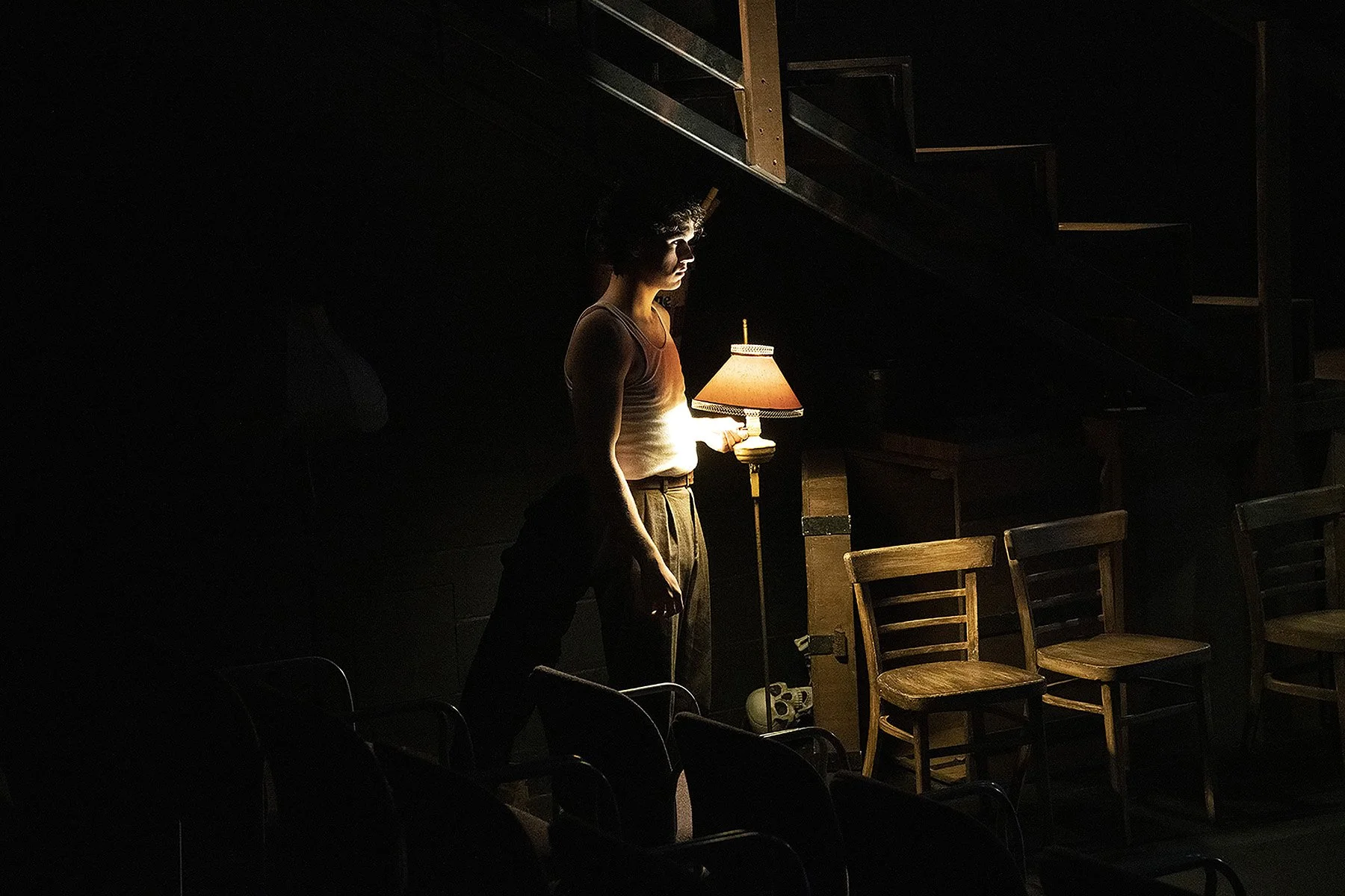 A woman standing in a dimly lit room, illuminated by a lamp she is holding, with empty chairs arranged behind her.
