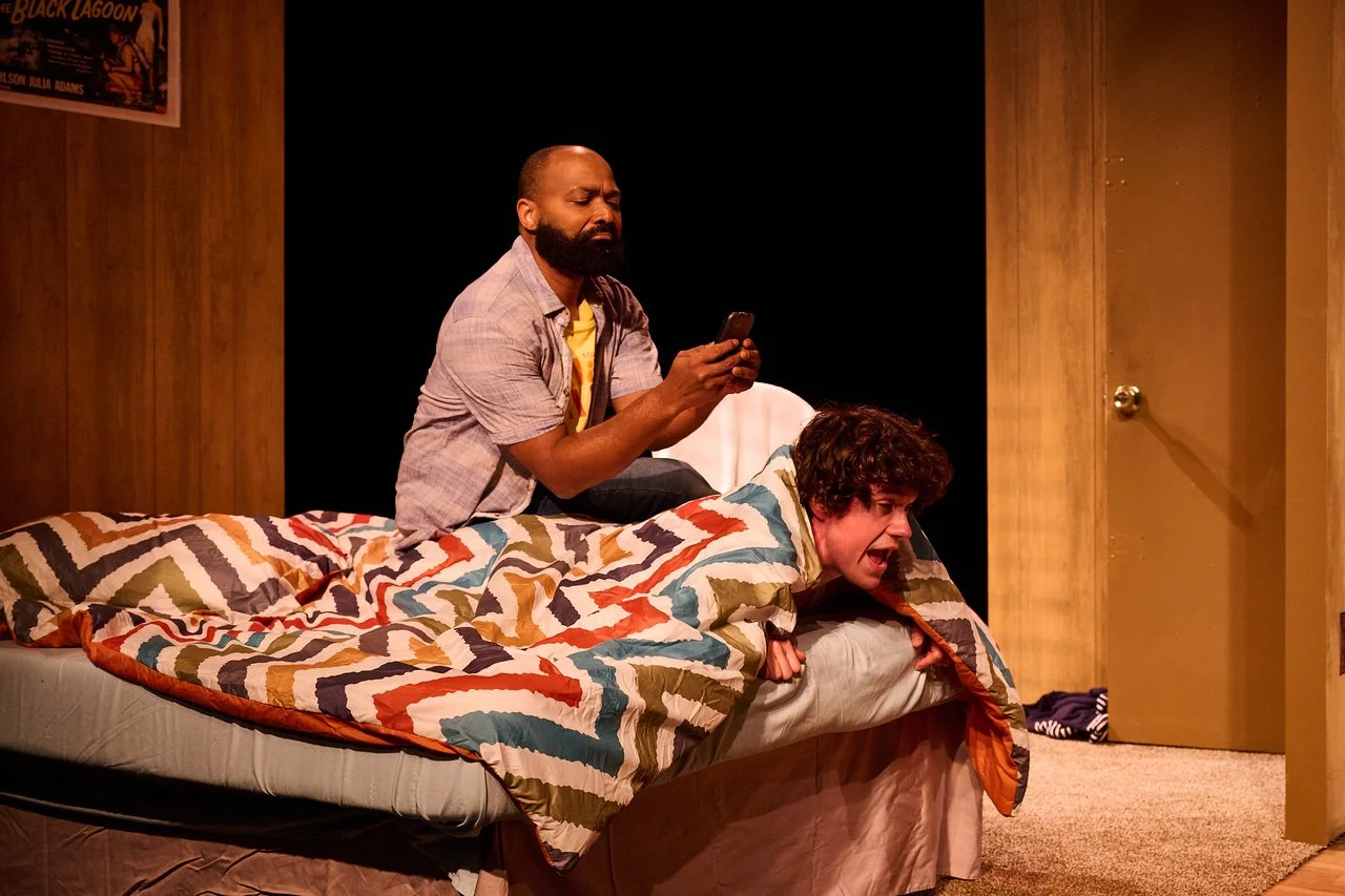 Two men on a bed, one using a smartphone while the other appears distressed, in a room with wood-paneled walls and a poster on the wall.