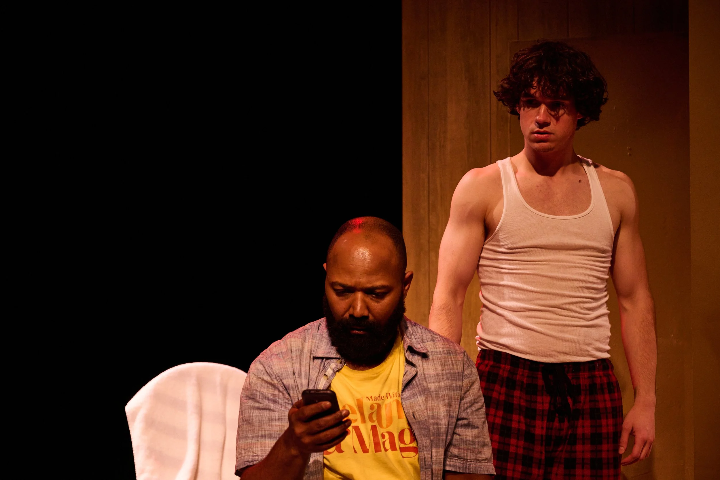Two men are seated, one is looking at his phone while the other stands behind him. The man standing has curly dark hair and is wearing a white tank top and plaid shorts. The seated man has a shaved head, a beard, and is wearing a checkered shirt over