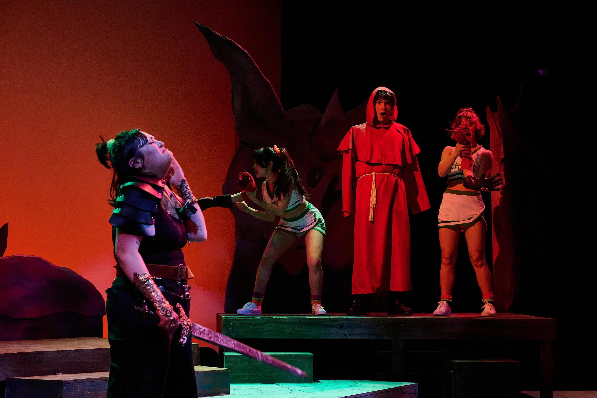 The image depicts a theatrical stage scene with five actors in colorful costumes. One actor is sitting in the foreground wearing a black and purple outfit with accessories and glasses, while three others are standing on a platform in the background, 