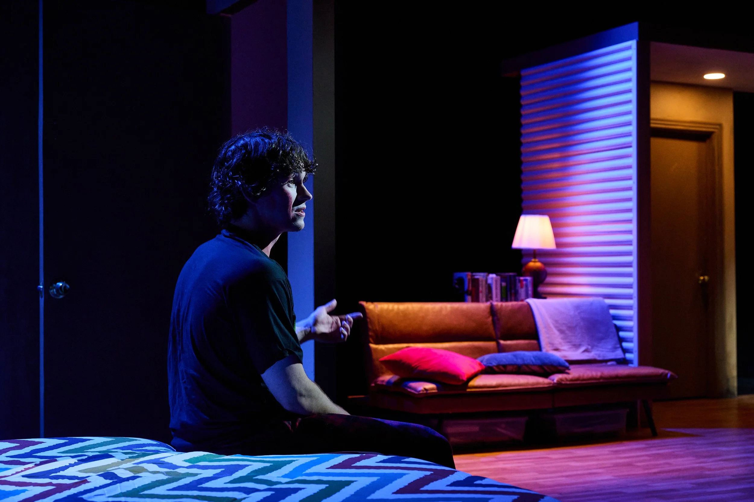 A man with short curly hair and a dark shirt sitting in a dimly lit room with neon lighting, facing sideways. There is a colorful couch with cushions, a bookshelf, a small lamp, and a textured wall with blue and purple lighting.