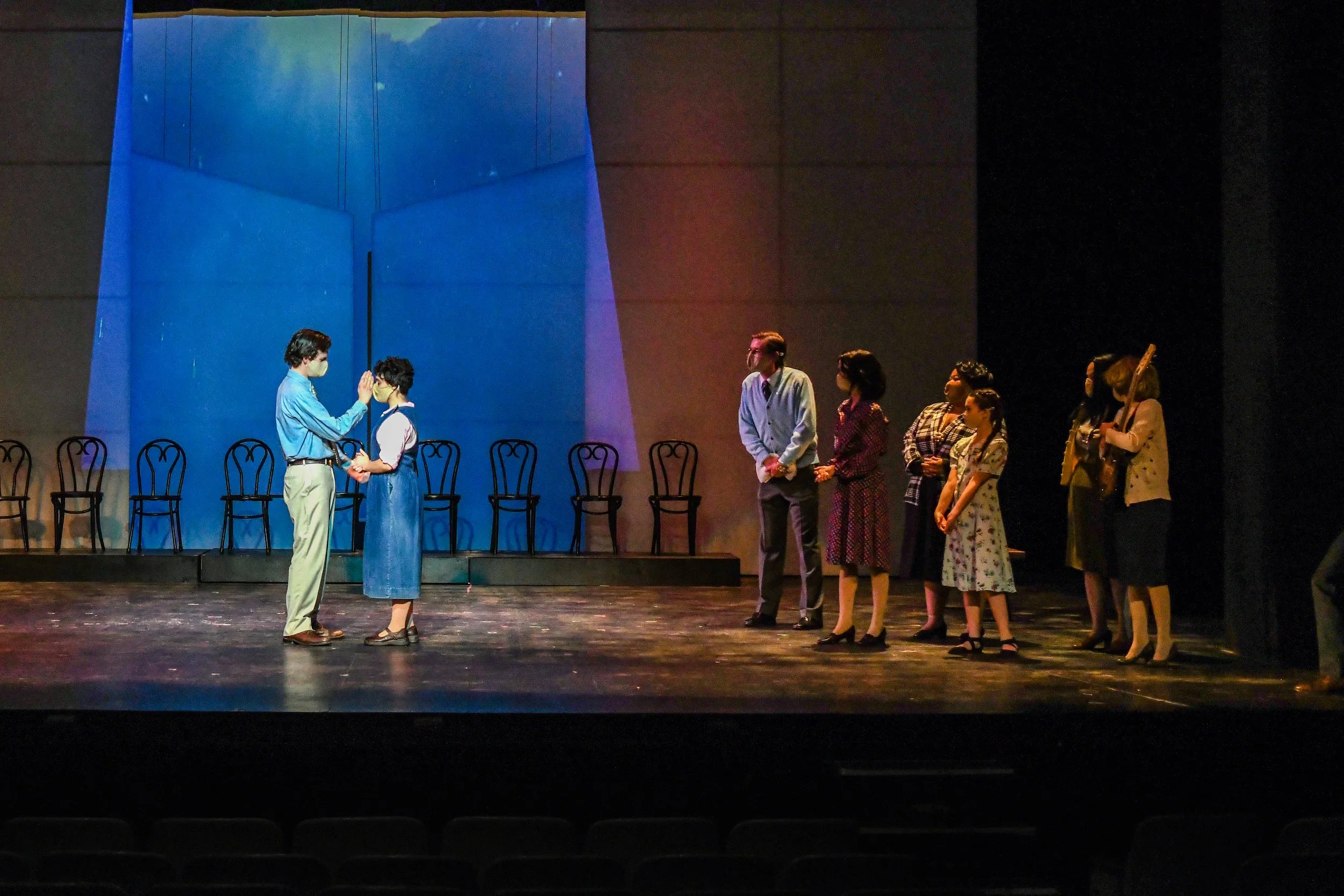 The image depicts a group of people standing on a stage, with a blue projection or light backdrop behind them. Two people in the foreground are engaging in a handshake or a close conversation, both wearing face masks. Several chairs are lined up agai