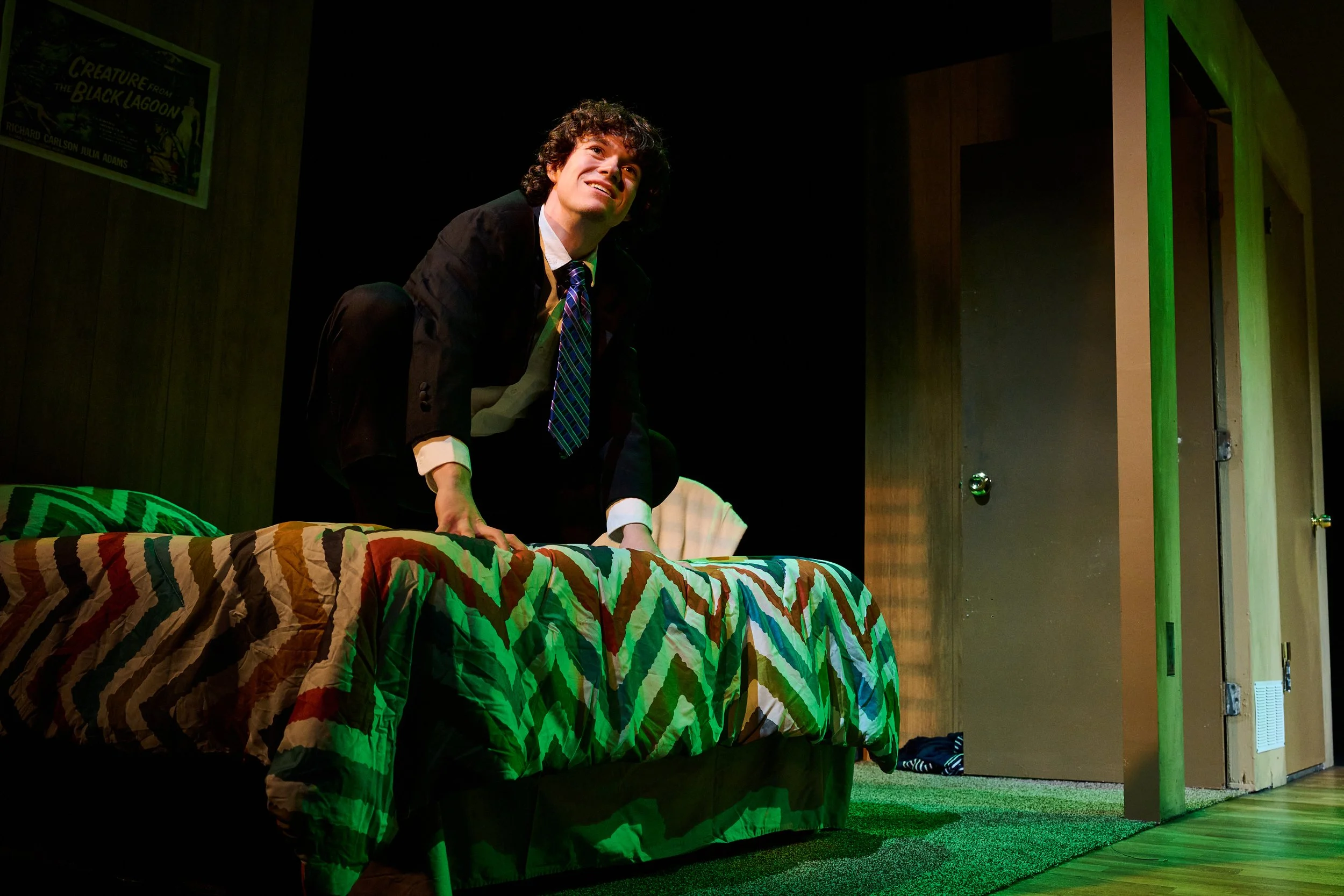 A young man with curly hair in a dark suit and colorful striped tie is squatting on a bed with a colorful zigzag patterned blanket, smiling and looking upward. The room has wooden walls, a poster on the wall, and a doorway with a green light illumina