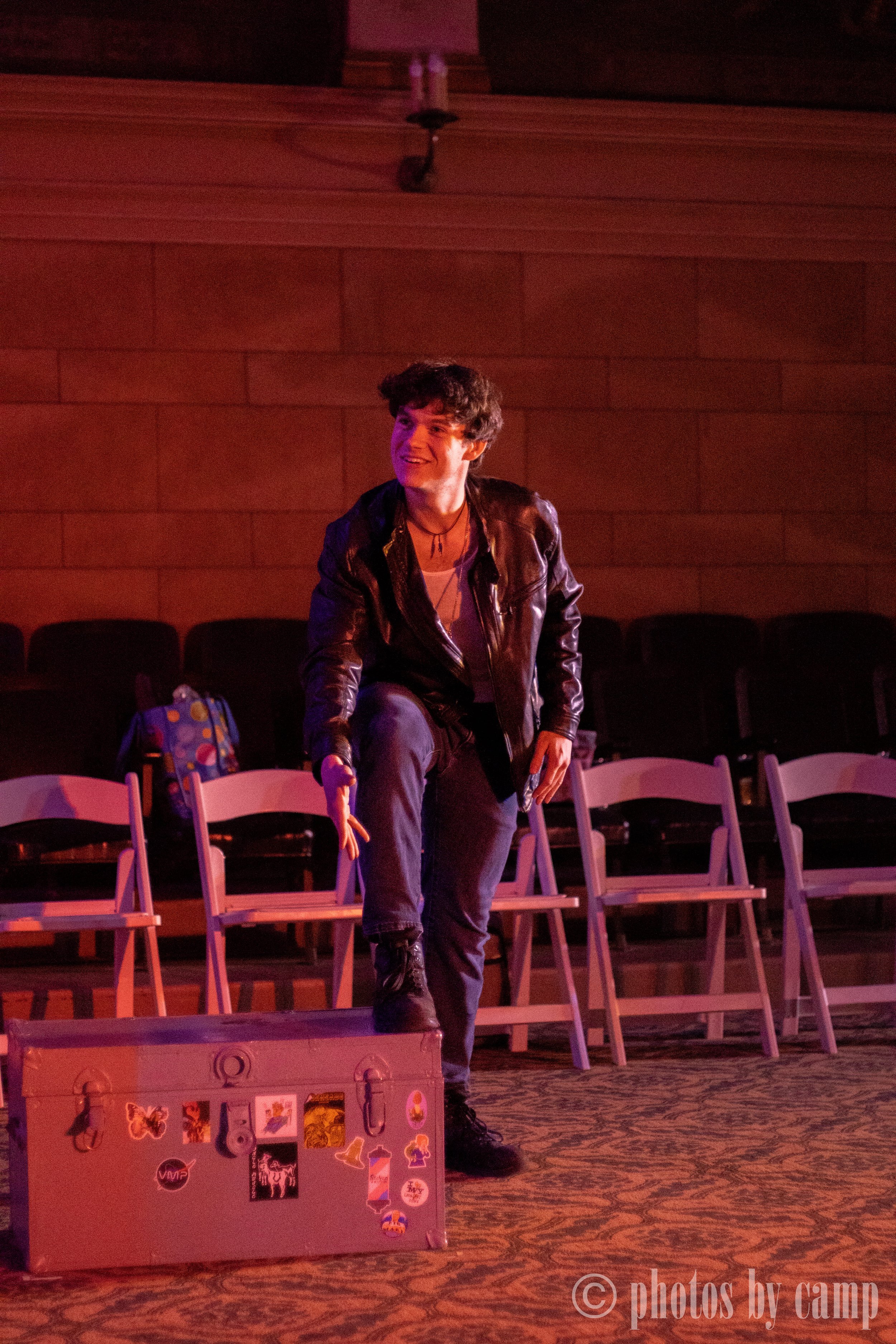 A woman with short curly hair wearing a leather jacket and gray pants, on a stage or performance space, surrounded by chairs and a box with various stickers, with a red lighting ambiance.